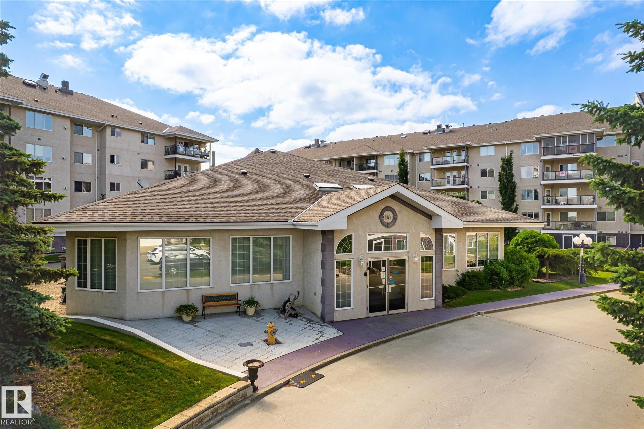 Community building entrance featuring a gabled roof, stucco exterior, and large windows - #310 237 Youville Dr E Nw, Edmonton, AB - Outdoor With Balcony