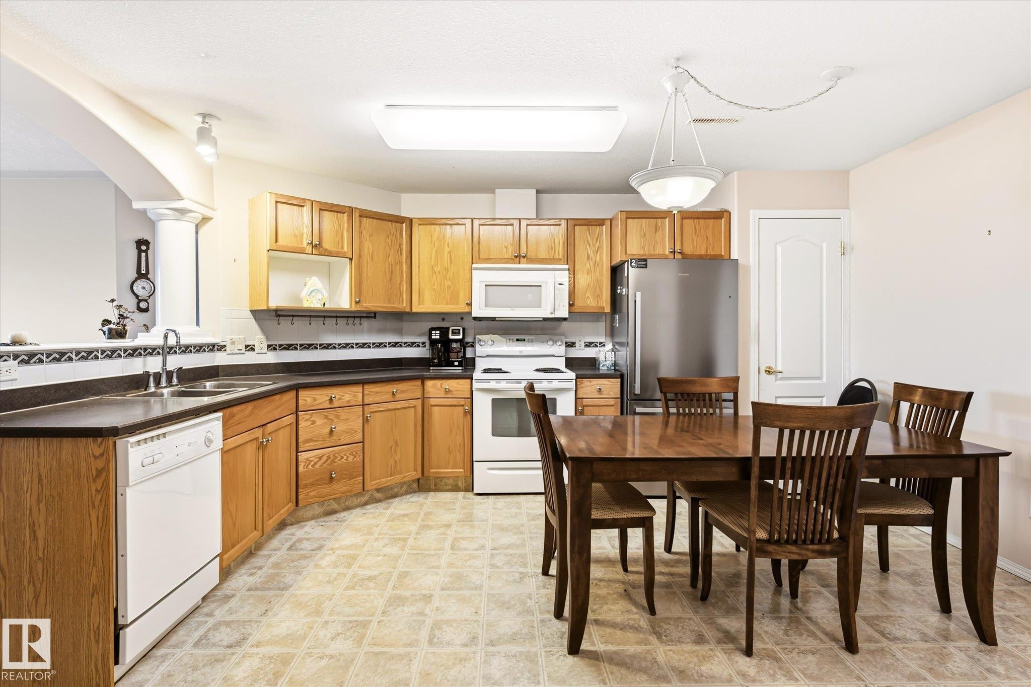 Open-concept kitchen featuring wood cabinetry, white appliances, and a stainless steel refrigerator - #310 237 Youville Dr E Nw, Edmonton, AB - Indoor Photo Showing Kitchen With Double Sink