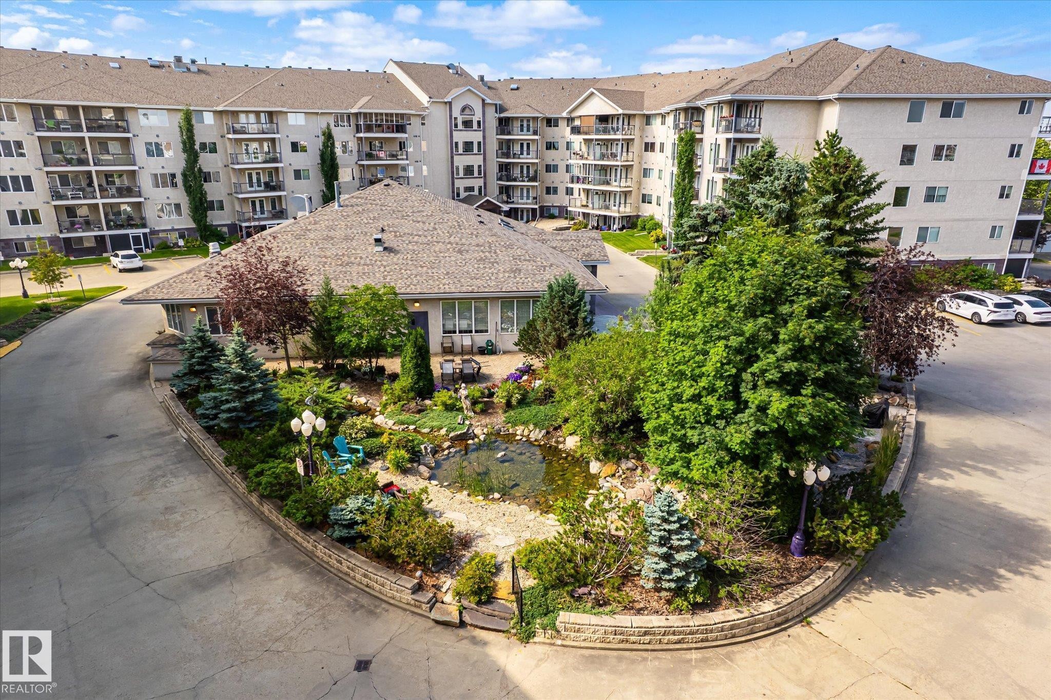 Expansive multi-story building featuring numerous balconies, a textured shingle roof, and a stone-edged landscaped garden with a central pond - #310 237 Youville Dr E Nw, Edmonton, AB - Outdoor With Balcony