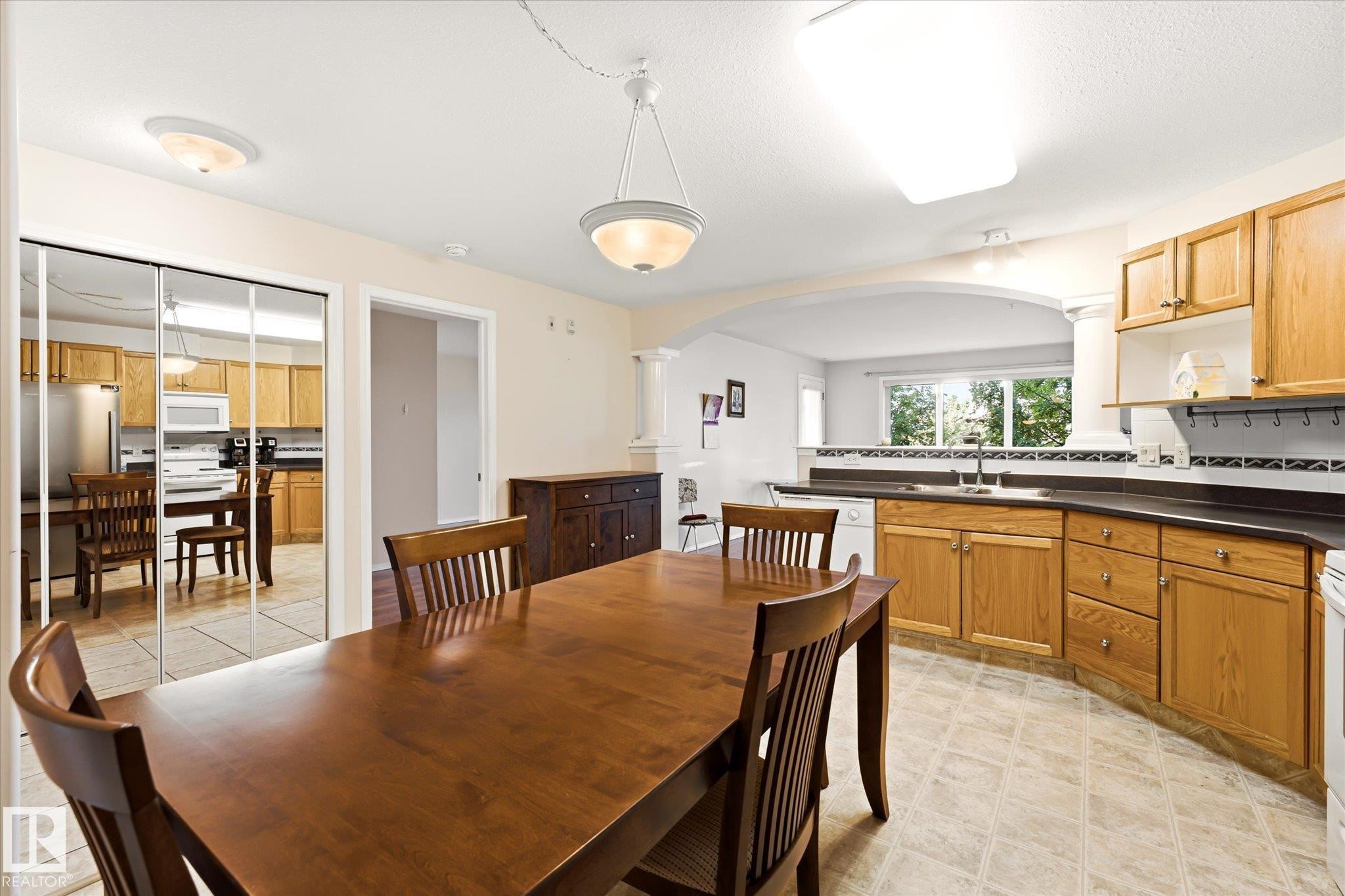 Dining area with wood-finish flooring and integrated kitchen featuring wood cabinetry, laminate countertops, and a window overlooking exterior greenery - #310 237 Youville Dr E Nw, Edmonton, AB - Indoor