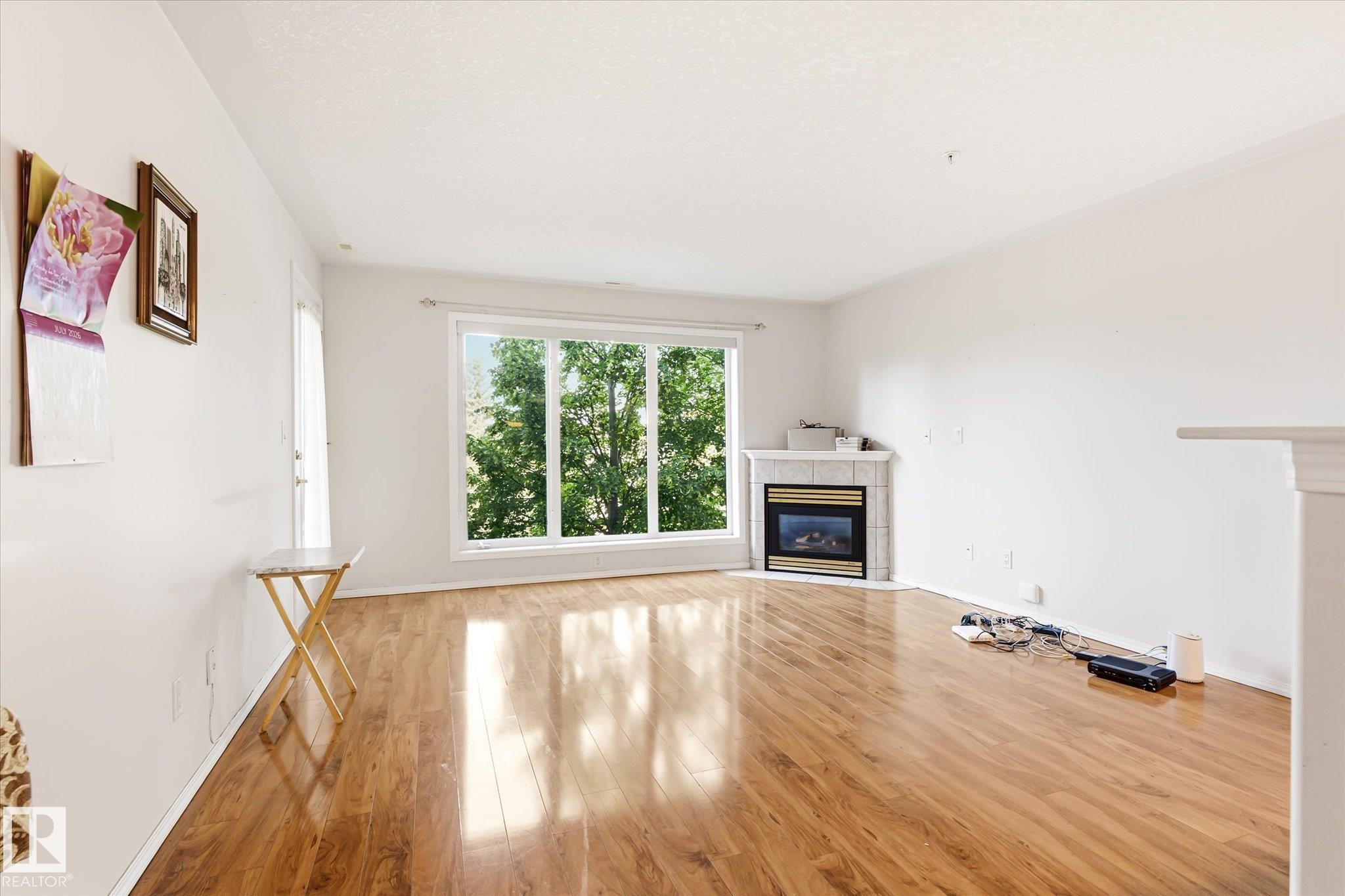 Spacious living area featuring expansive wood-finish flooring, a corner-situated fireplace with a tile surround, and a large bay window offering abundant natural light - #310 237 Youville Dr E Nw, Edmonton, AB - Indoor Photo Showing Other Room With Fireplace