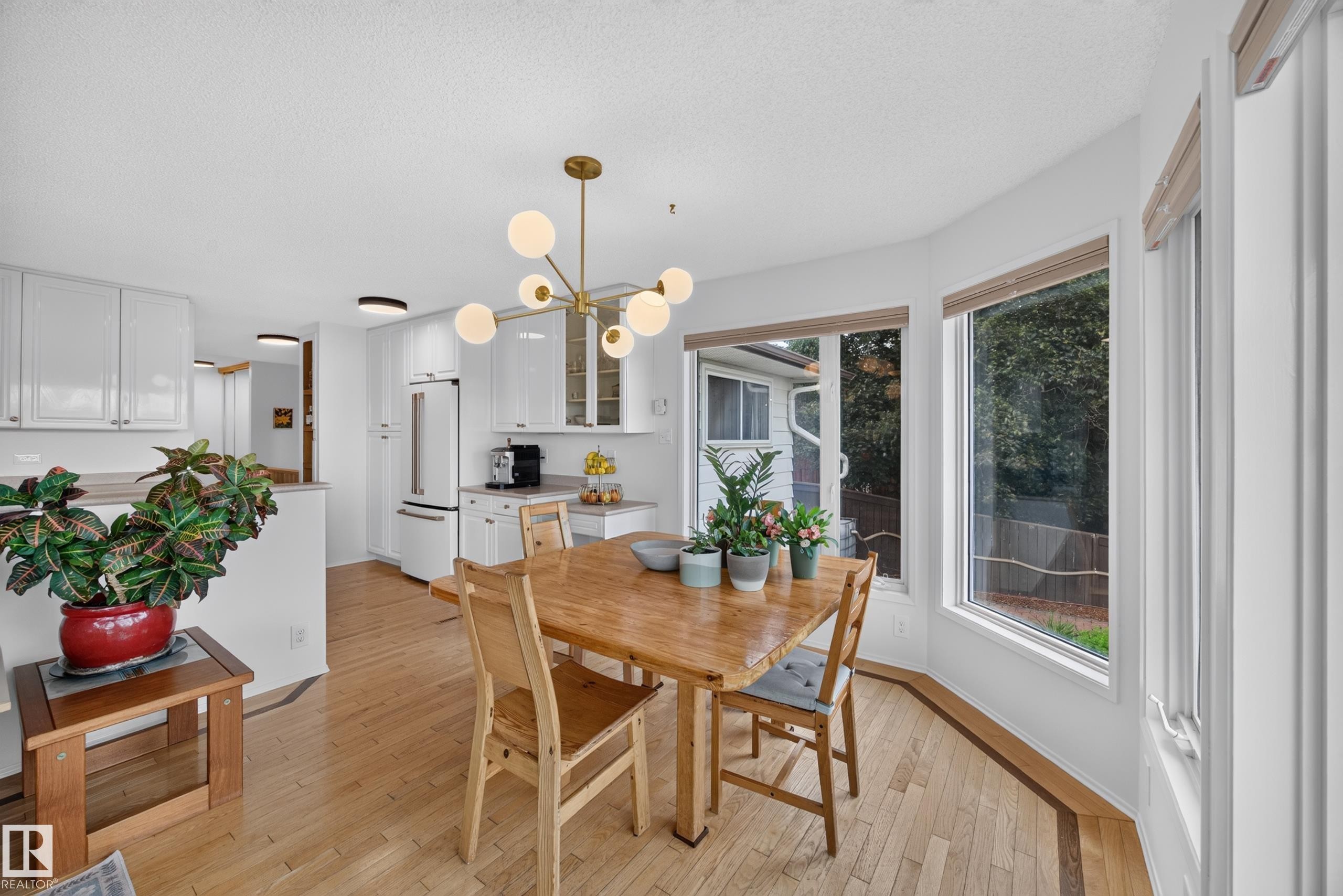 Spacious dining area featuring a modern brass chandelier, bay windows, wood-finish flooring, and white cabinetry - 3411 103 Avenue Nw, Edmonton, AB