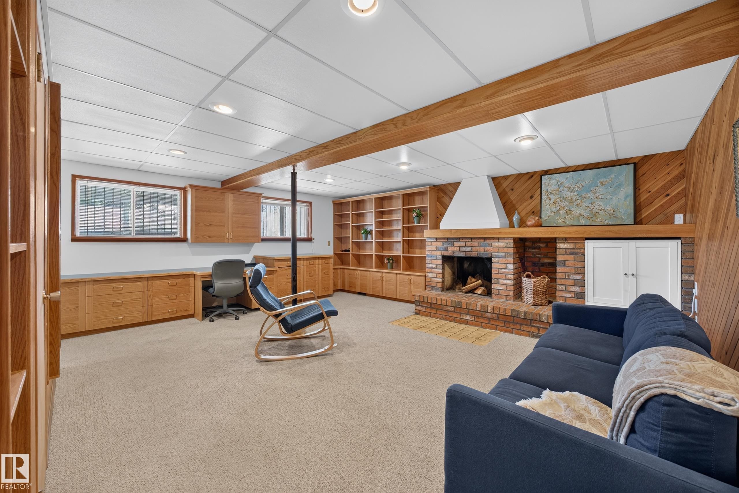 Spacious lower-level room featuring a brick fireplace with a wood mantel, extensive built-in shelving, and custom cabinetry with integrated desk space - 3411 103 Avenue Nw, Edmonton, AB
