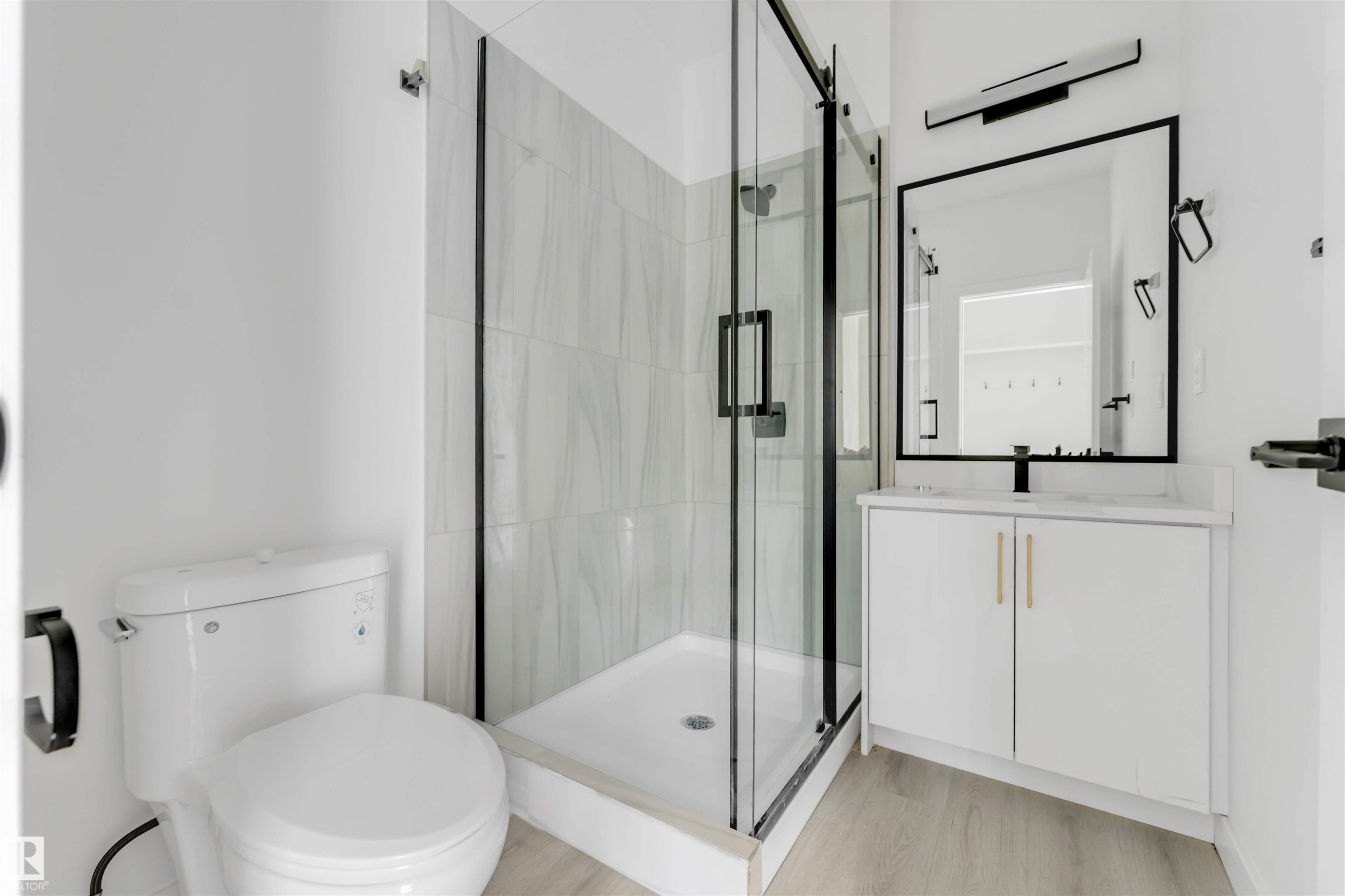 Bathroom featuring a glass-enclosed shower with tiled walls, a white vanity with gold-tone hardware, a rectangular framed mirror, and light wood-finish flooring - 3605 112 Ave, Edmonton, AB
