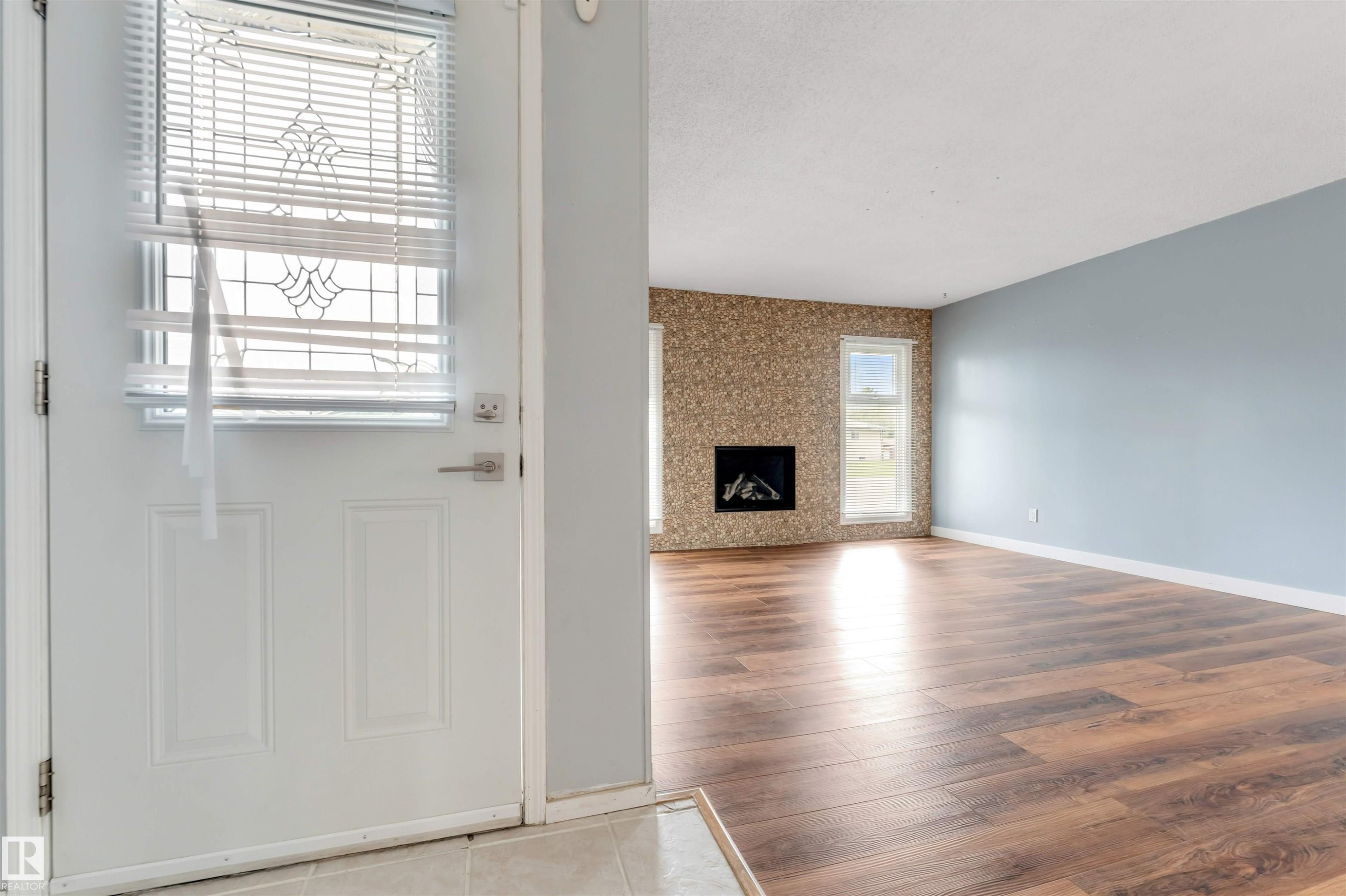 Entryway with a paneled door featuring decorative glass inserts, opening to an interior space with wood-finish flooring, a contemporary fireplace with a stone-finish surround, and a light blue painted wall - 193 Woodvale Road W, Edmonton, AB