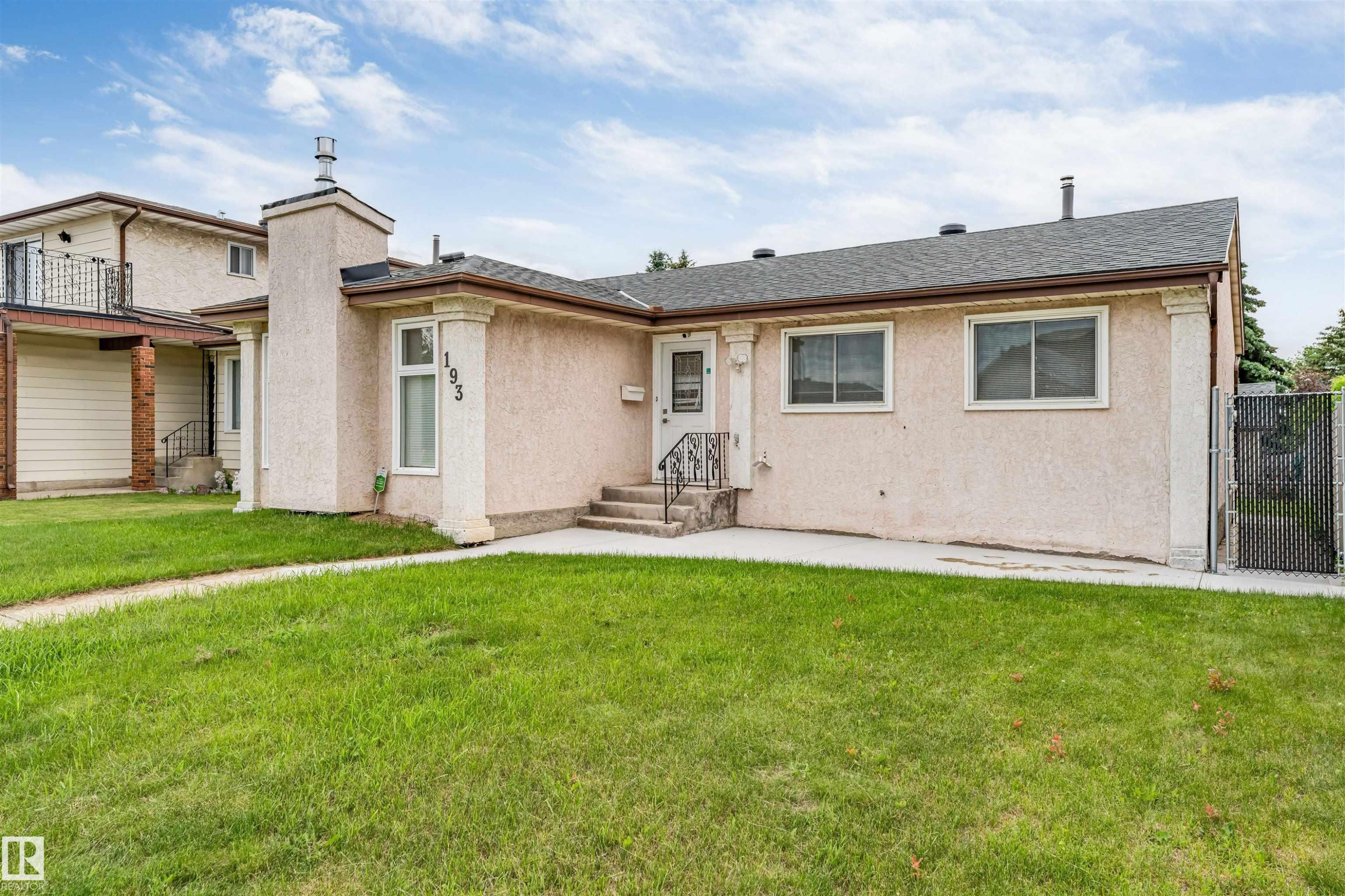 Stucco exterior with a peaked roofline, featuring a front porch with railing, a prominent chimney, and a chain-link fence at the property edge - 193 Woodvale Road W, Edmonton, AB