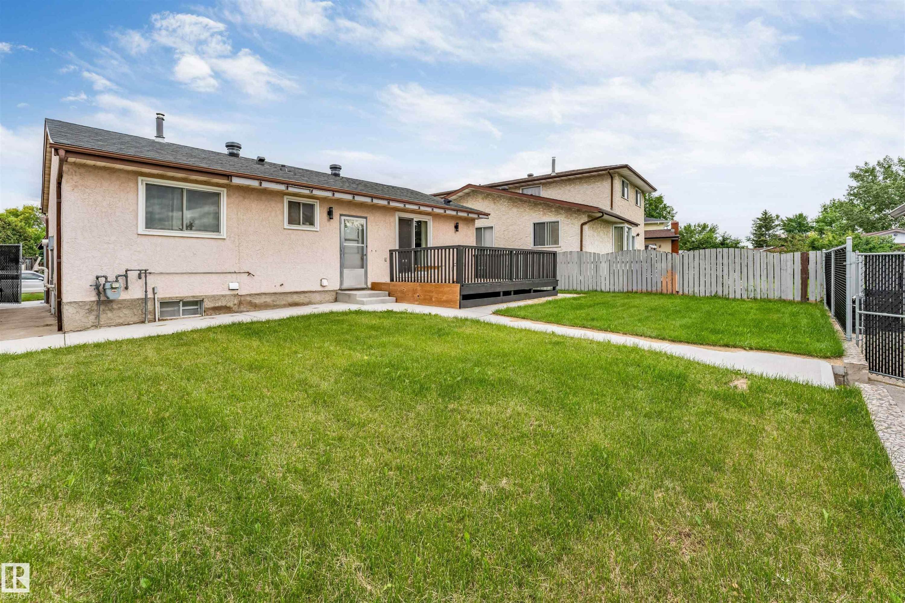 Expansive rear yard featuring green lawn, a concrete pathway, a dark wood-finish deck with railings, and a chain-link fence with a gate - 193 Woodvale Road W, Edmonton, AB