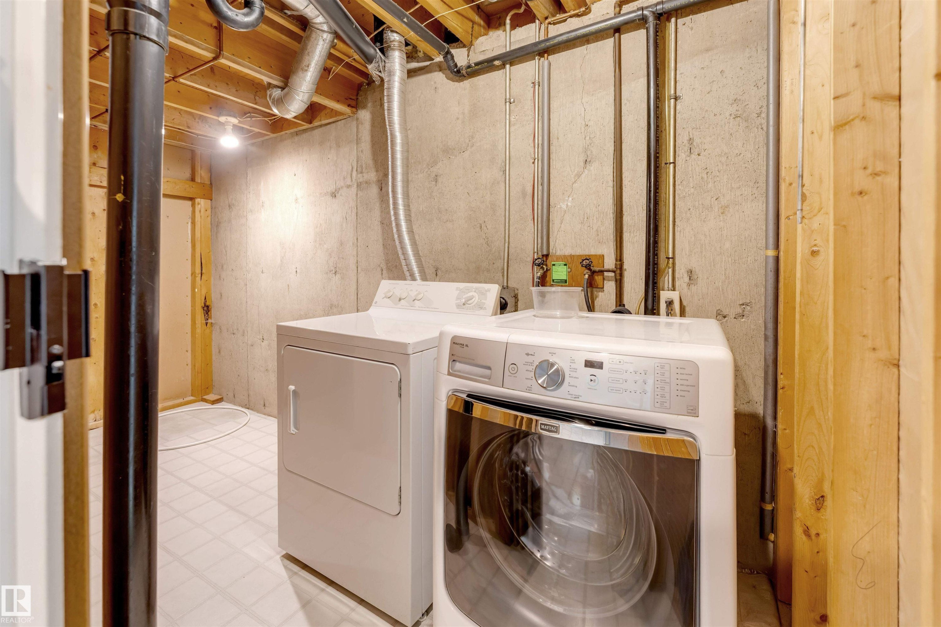 Basement utility area featuring a washer and dryer, exposed concrete walls, wood ceiling joists, and white patterned flooring - 193 Woodvale Road W, Edmonton, AB