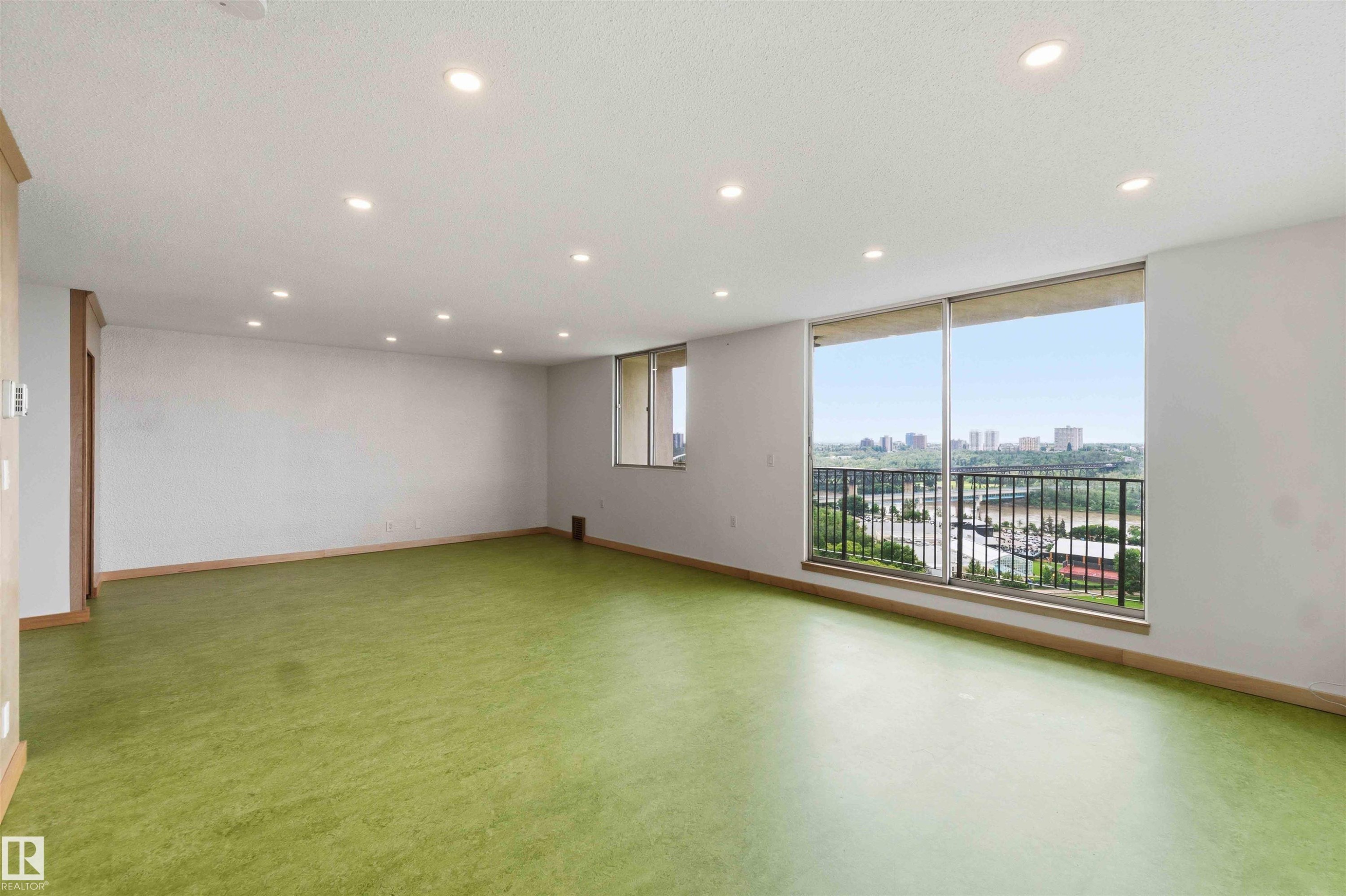 Expansive interior space featuring a full-wall window with a metal railing, recessed ceiling lighting, and a panoramic cityscape - #1008 11307 99 Av Nw, Edmonton, AB