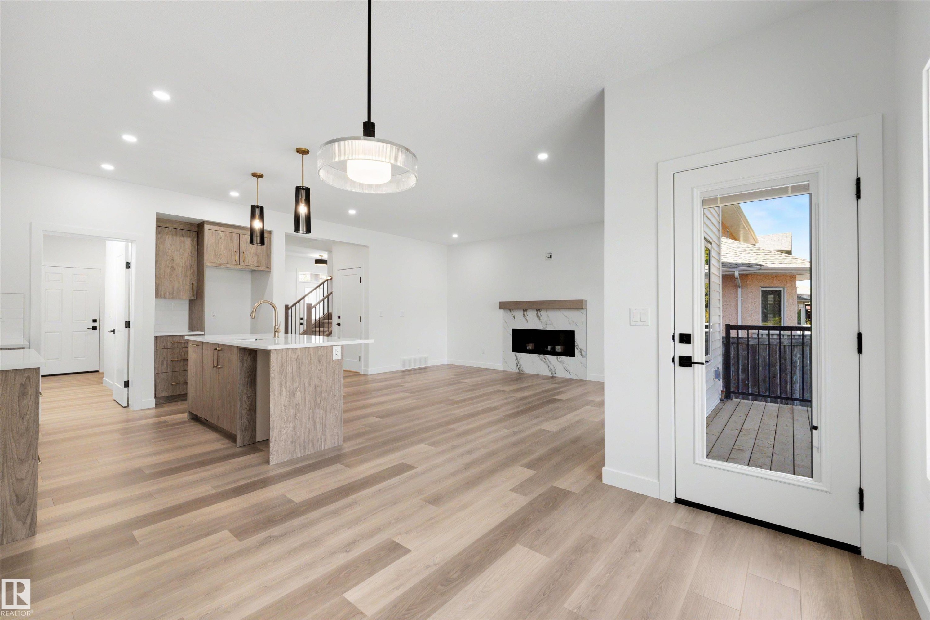Open-concept main living area featuring wood-finish flooring throughout, a modern kitchen island with a quartz countertop, recessed lighting, a contemporary fireplace with a stone surround, and a glass-paneled door leading to an outdoor deck - 30 Elsinore Place Nw, Edmonton, AB