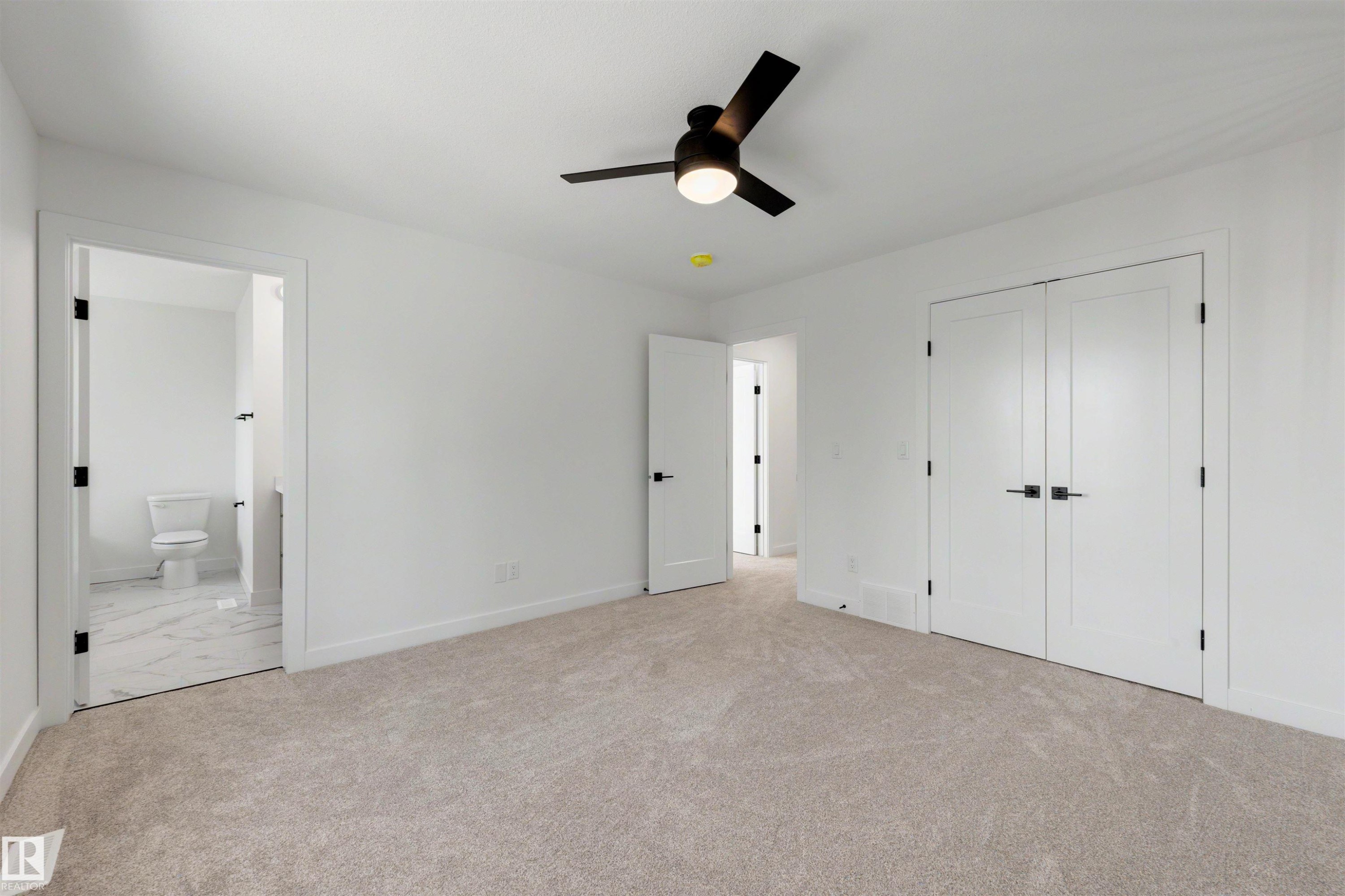 Spacious carpeted room featuring a ceiling fan with integrated lighting, dual white panel doors with matte black hardware, and an en-suite bathroom with marble-look tile flooring - 30 Elsinore Place Nw, Edmonton, AB