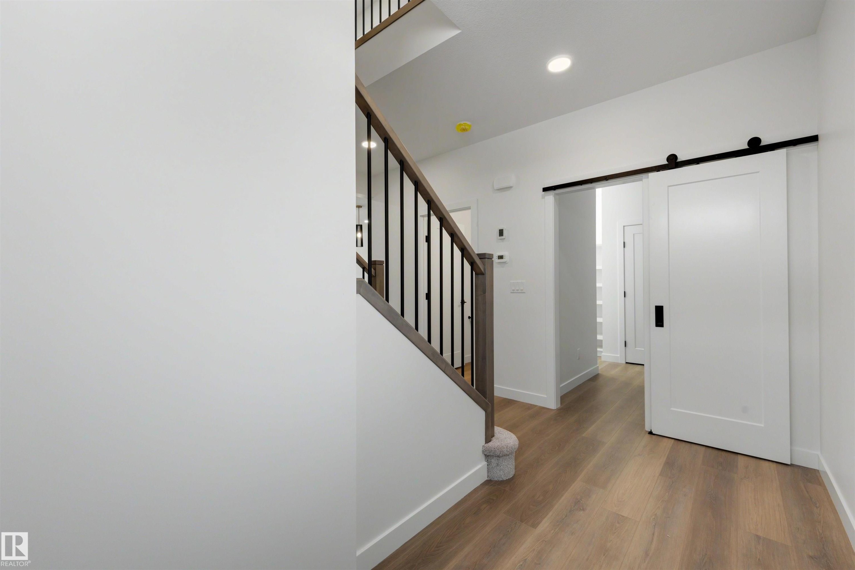 Modern interior hallway featuring wood-finish flooring, a contemporary staircase with dark metal balusters, and a sliding barn door with black hardware - 30 Elsinore Place Nw, Edmonton, AB