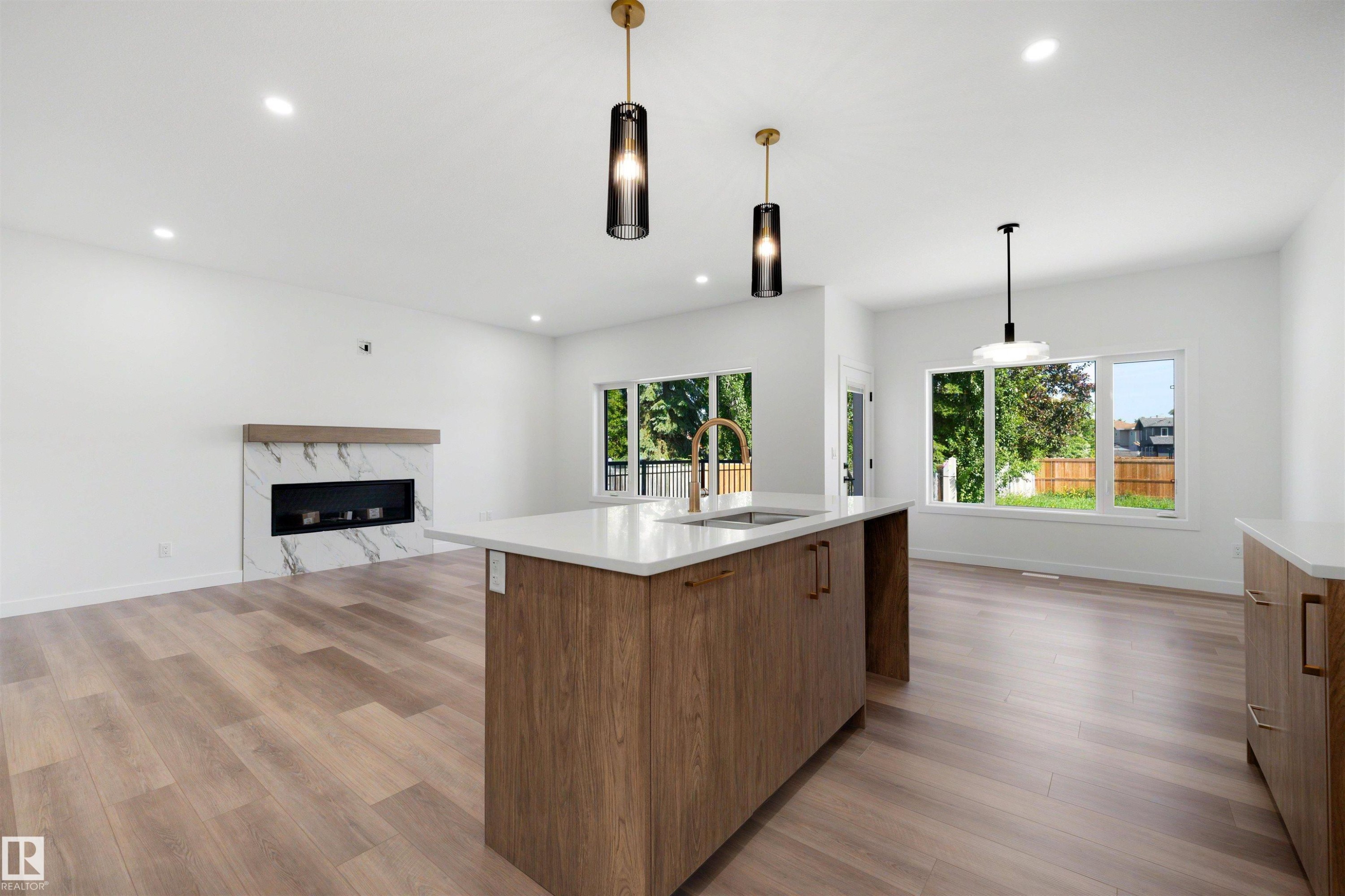 Open-concept living area featuring a kitchen island with a gold-tone faucet, wood-finish flooring, a modern fireplace with a marble-patterned surround, recessed lighting, and large windows - 30 Elsinore Place Nw, Edmonton, AB