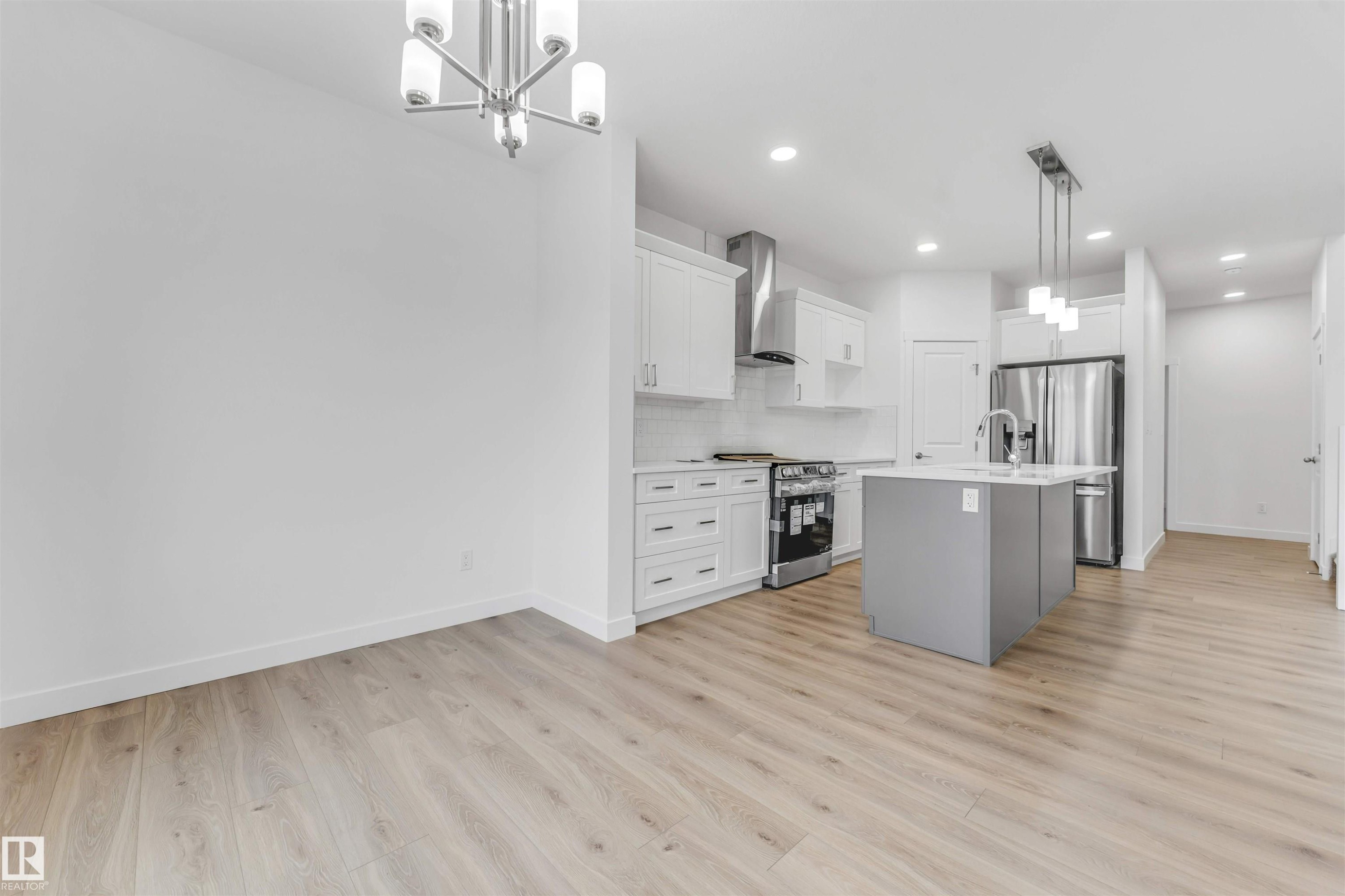 Modern kitchen featuring a gray island with integrated sink, stainless steel appliances, white cabinetry, and light wood-finish flooring throughout - 1972 210 Street, Edmonton, AB