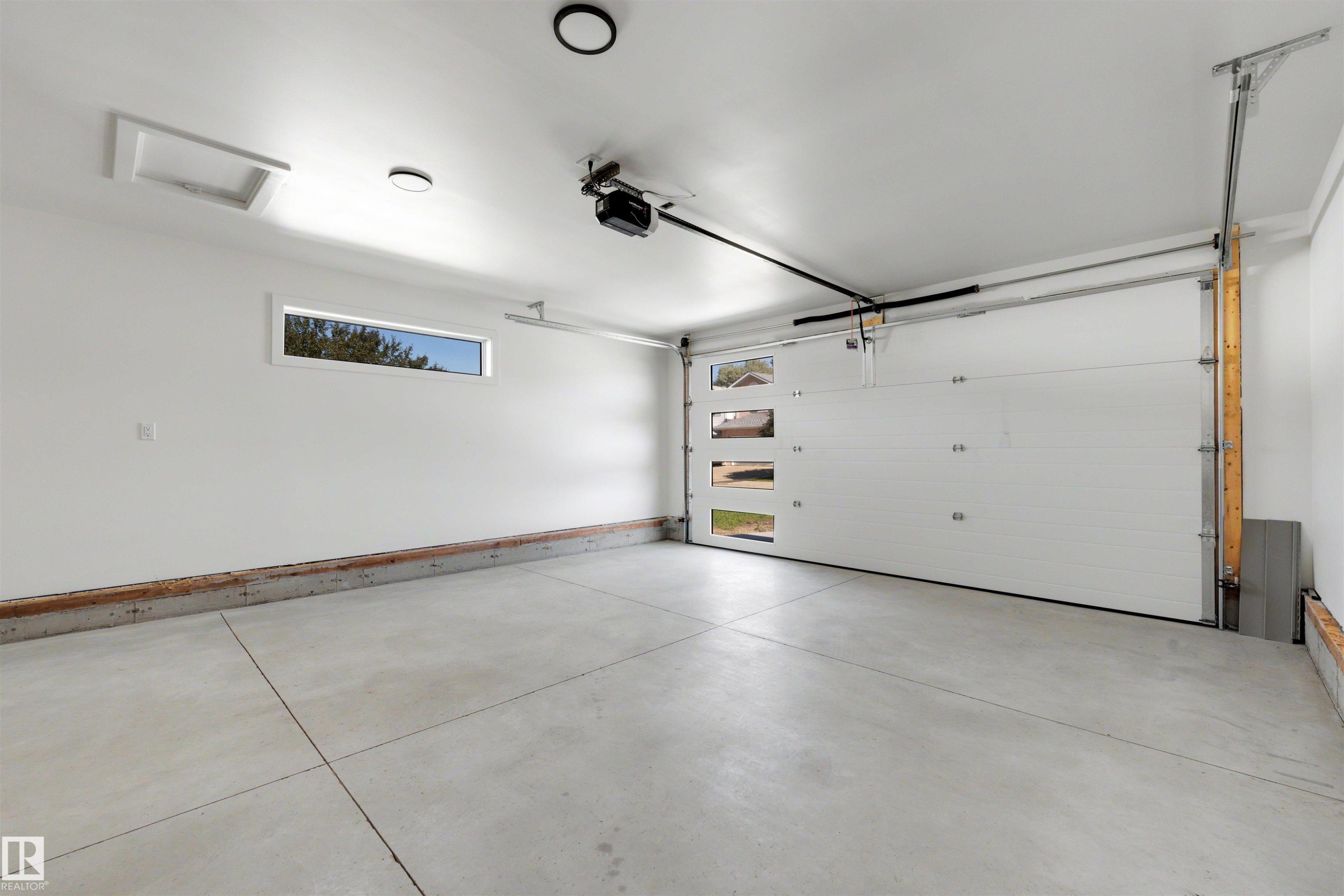 Spacious garage featuring a concrete floor, white walls, modern overhead lighting, and an automatic garage door with rectangular window inserts - 39 Elsinore Place, Edmonton, AB