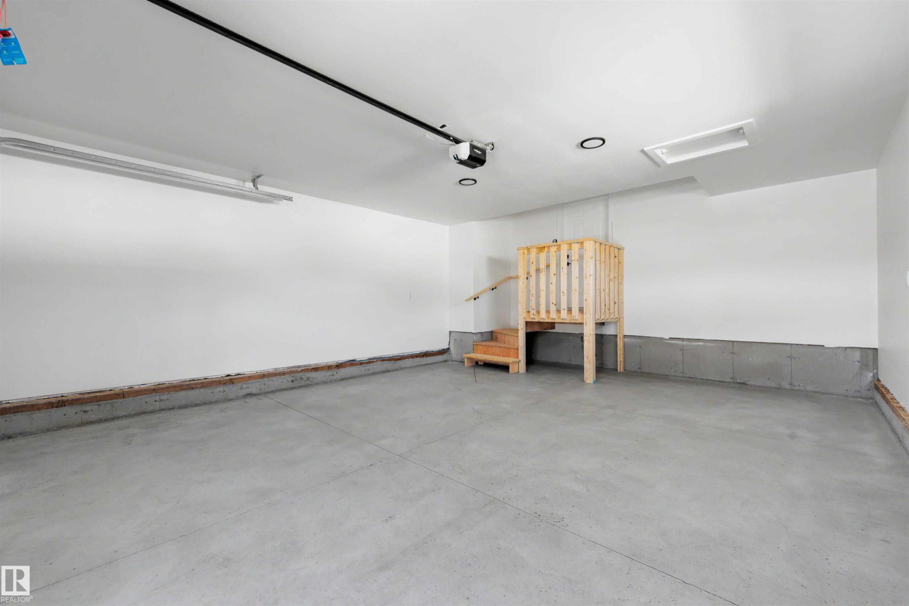 Spacious garage featuring a concrete floor, painted white walls, overhead lighting, an automatic garage door opener, and an elevated wooden platform with stairs and railing - 39 Elsinore Place, Edmonton, AB