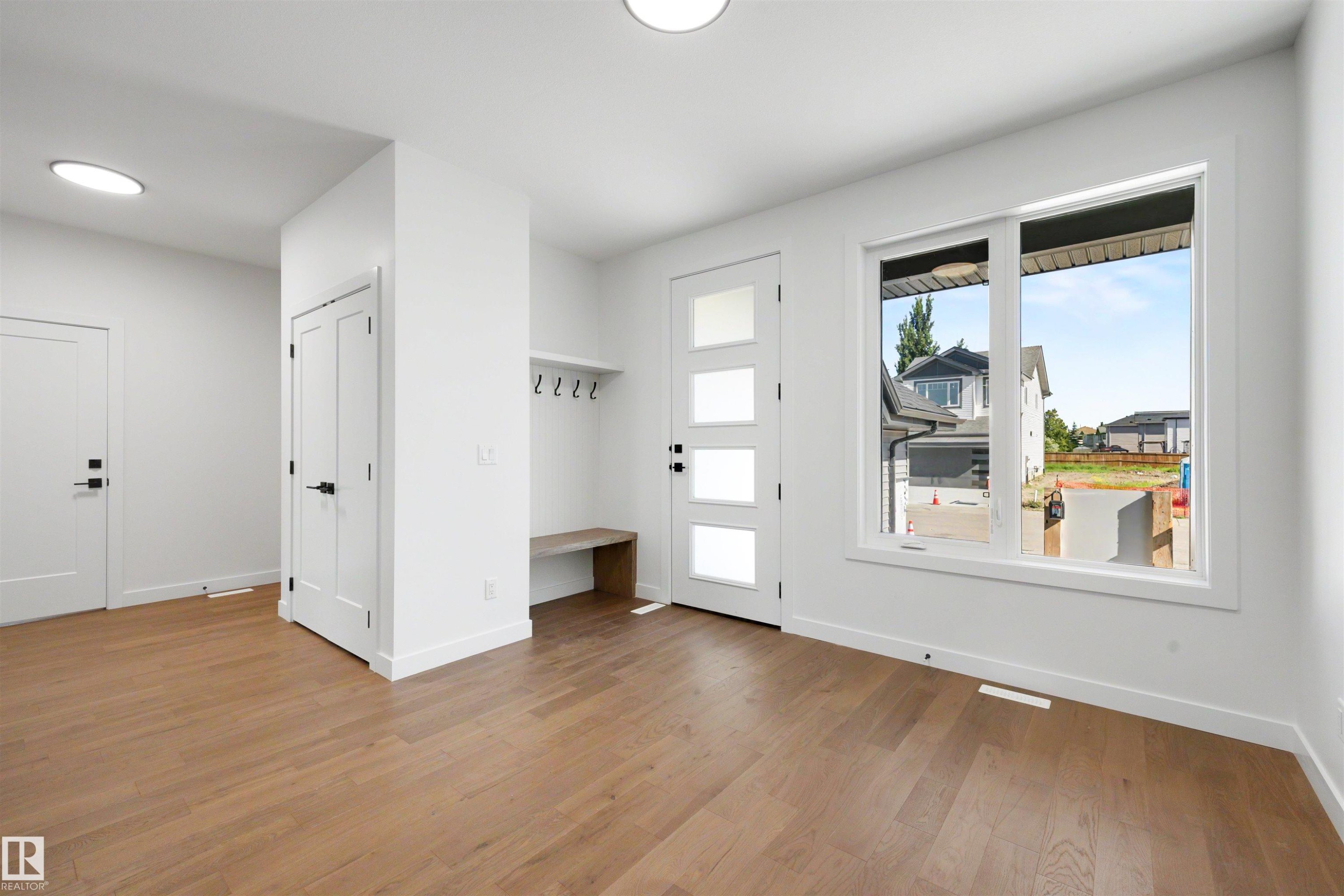 Entryway featuring wood-finish flooring, a modern front door with frosted glass panels, a built-in bench with coat hooks, crisp white walls, and a large window providing ambient light - 39 Elsinore Place, Edmonton, AB
