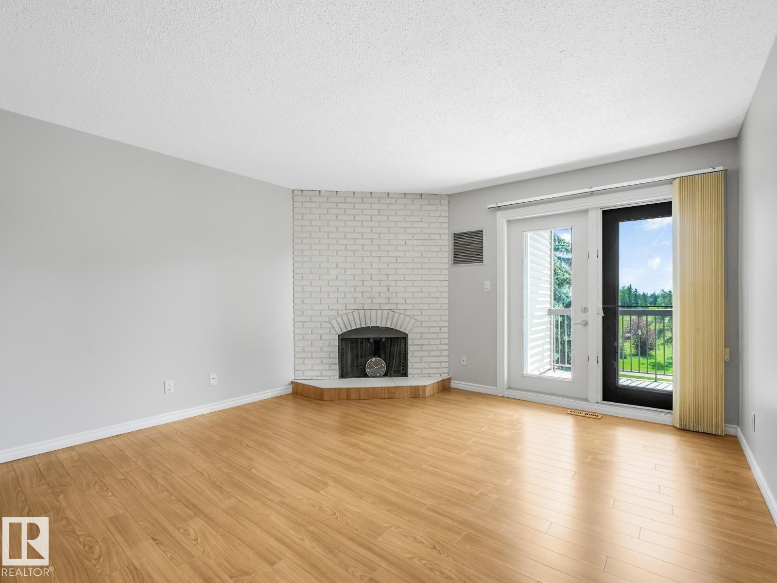 Spacious room featuring wood-finish flooring, a white brick fireplace, and light gray walls - #301 10528 29 Av Nw, Edmonton, AB