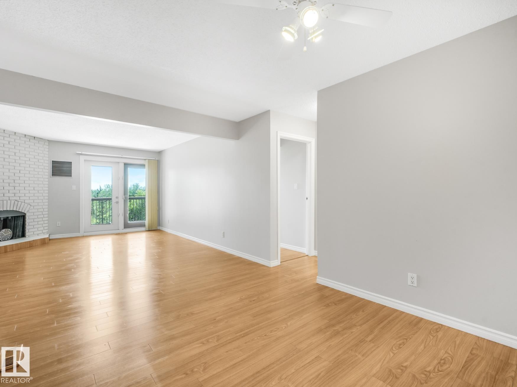 Open-concept living area featuring wood-finish flooring, a white brick fireplace, and dual French doors with a black metal railing - #301 10528 29 Av Nw, Edmonton, AB