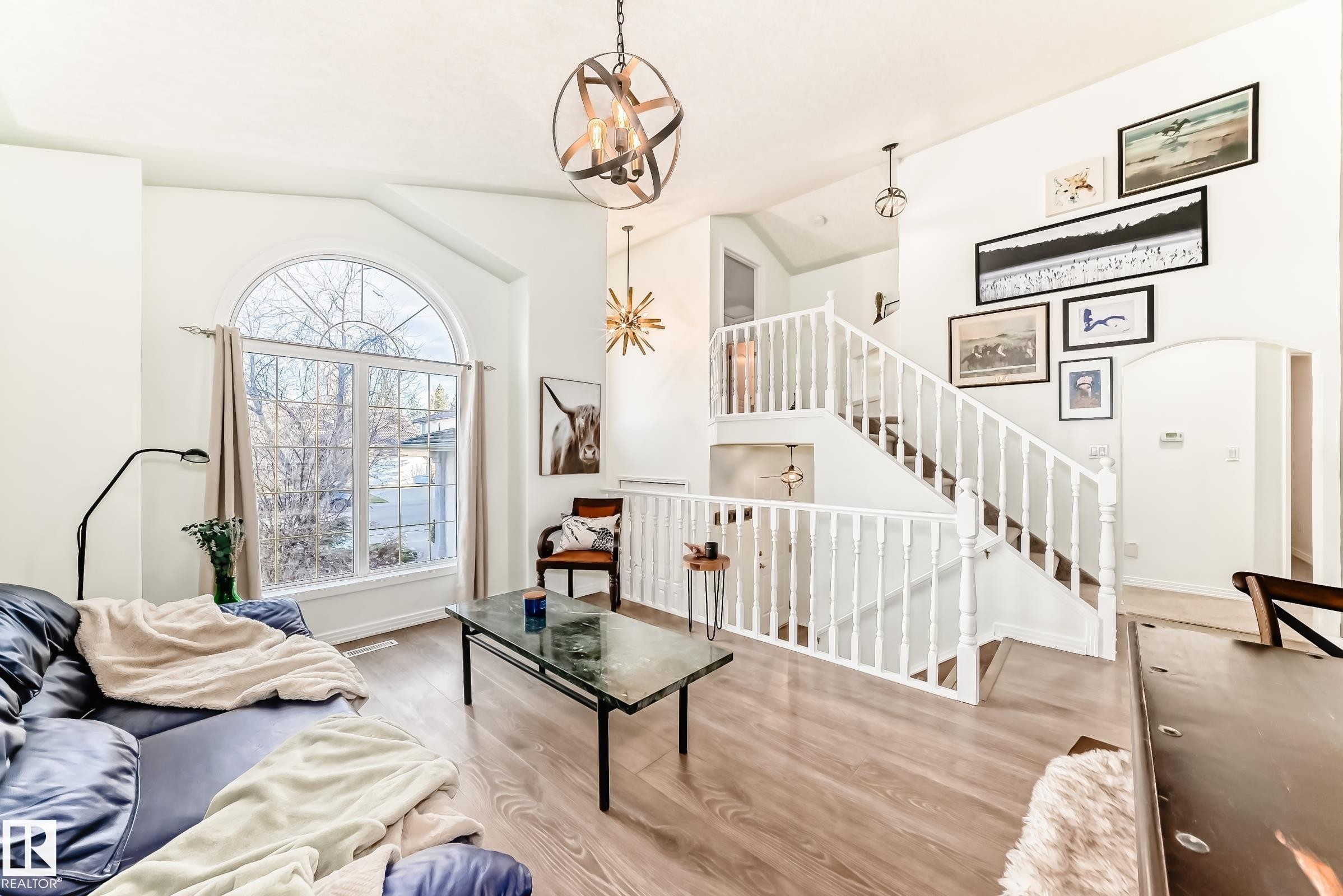 Vaulted ceiling living room featuring a large arched window, wood-finish flooring, a multi-level staircase with white balusters, and a tiered orb chandelier - 1619 Welbourn Cove, Edmonton, AB