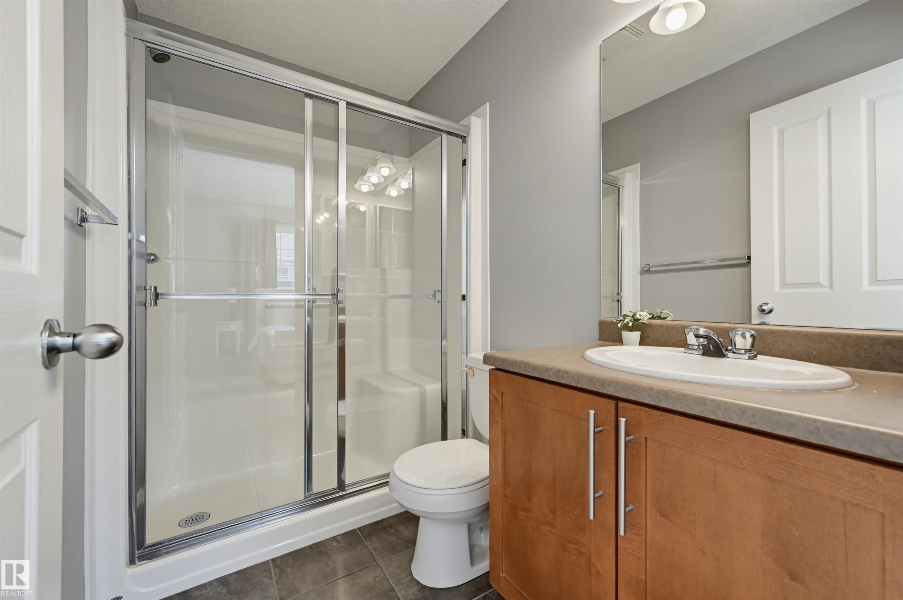 Shower enclosure with sliding glass doors, a single vanity with an integrated sink, wood-finish cabinetry, a neutral countertop, and a wall-mounted mirror - 126 2051 Towne Centre Boulevard, Edmonton, AB