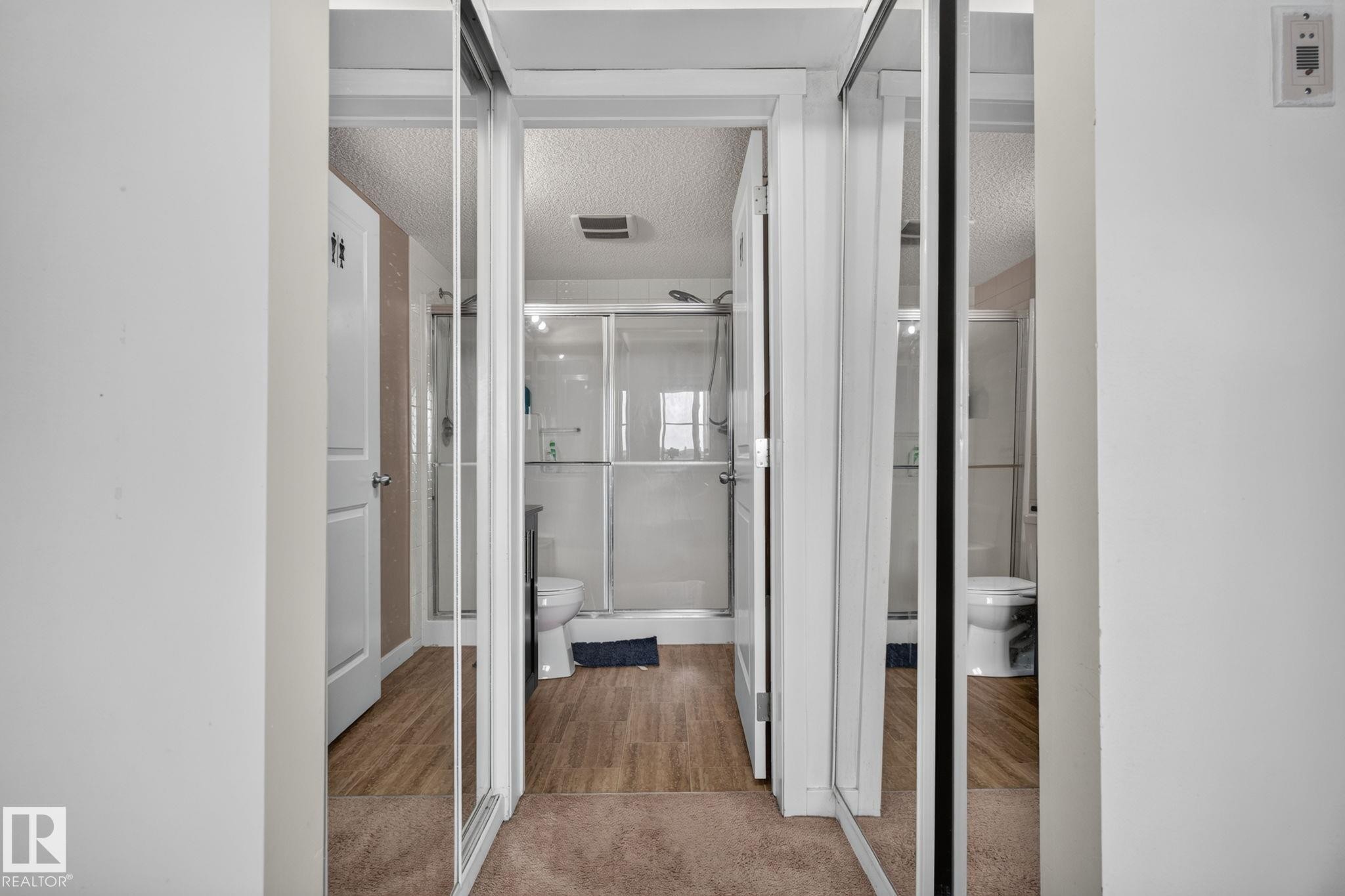 Sliding mirrored closet doors reflecting a bathroom with wood-finish flooring, a glass-enclosed shower, and a white toilet - 415 111 Watt Common, Edmonton, AB