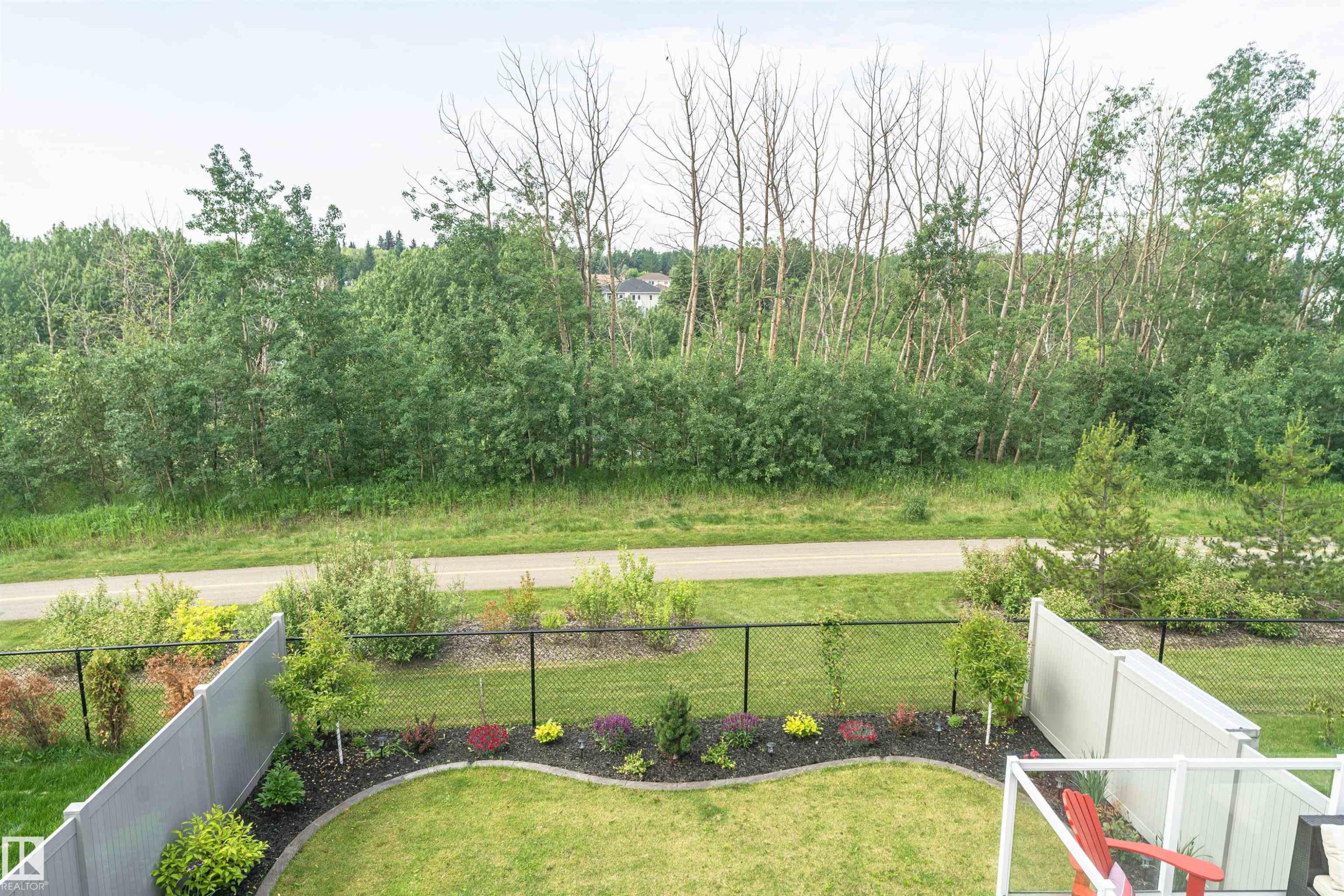 Expansive backyard featuring a manicured lawn, established garden beds with various plantings, and a black chain-link fence bordering a paved pathway and extensive natural foliage - 102 Horton Way, Ardrossan, AB