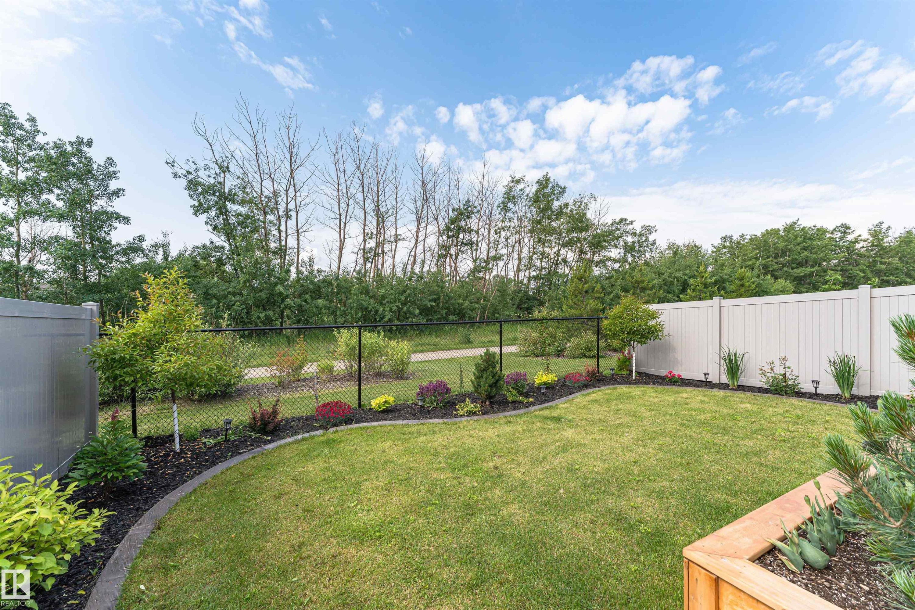 Expansive backyard featuring a manicured lawn, established garden beds with diverse plantings, a black chain-link fence, and a white privacy fence - 102 Horton Way, Ardrossan, AB