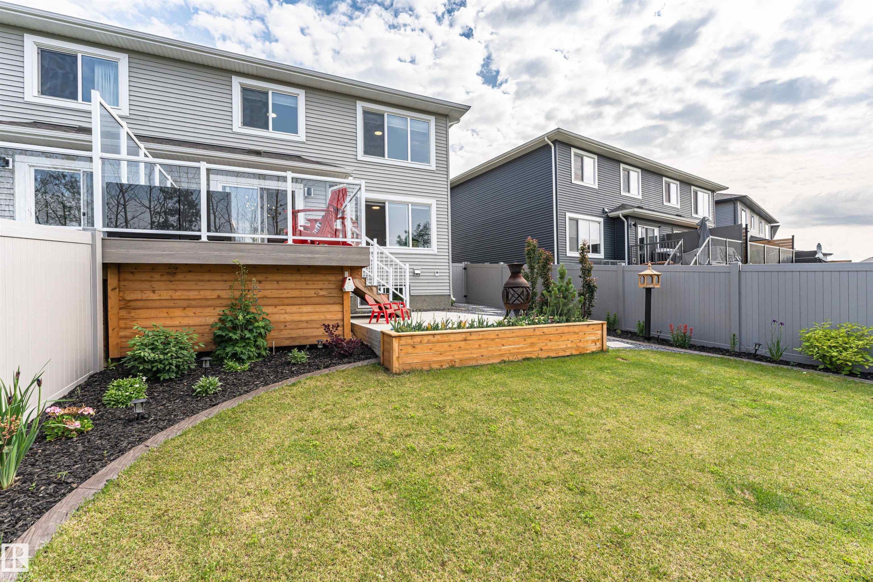 Expansive backyard featuring a manicured lawn, a raised wooden deck with glass panel railings, and a lower patio area - 102 Horton Way, Ardrossan, AB