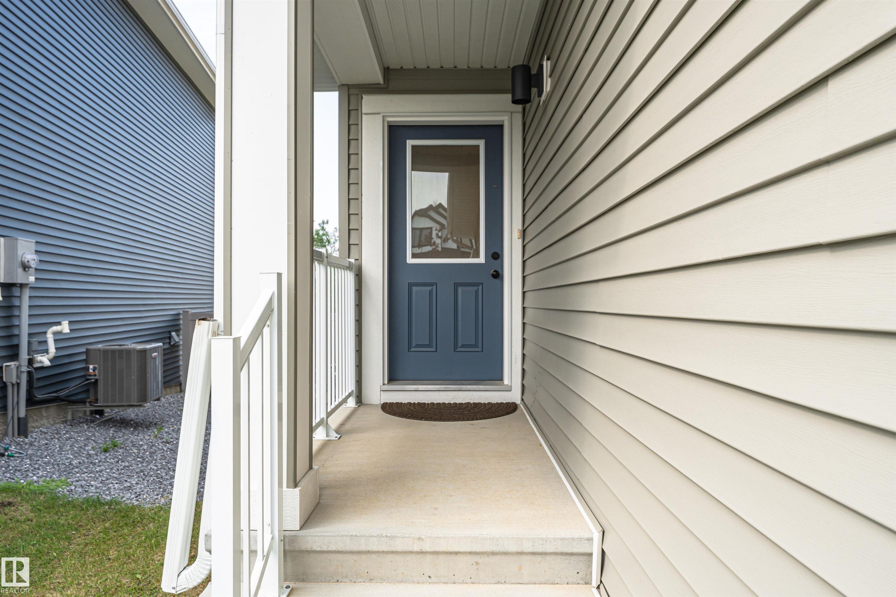 Entryway featuring a blue paneled door with an upper glass insert, bordered by light-colored siding and a concrete porch with white railings - 102 Horton Way, Ardrossan, AB