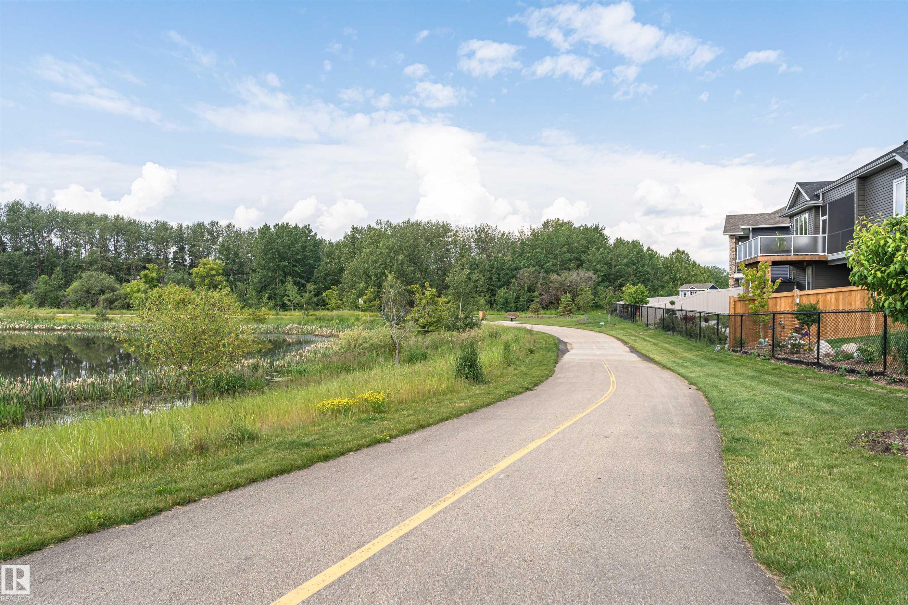 Paved pathway alongside a naturalized pond, bordered by manicured lawn and mature trees, with modern homes featuring balconies and privacy fencing visible in the background - 102 Horton Way, Ardrossan, AB