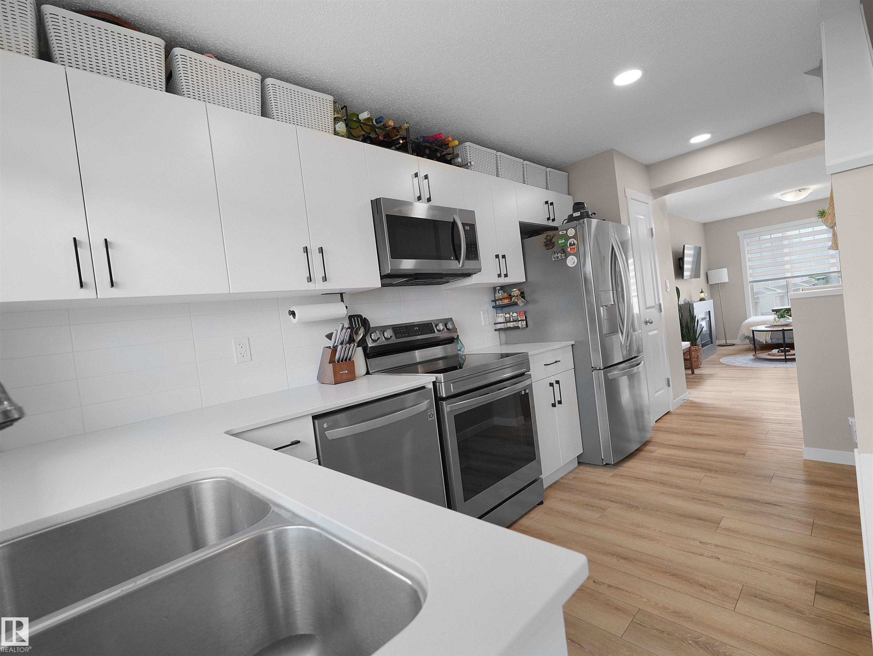 Contemporary kitchen featuring white cabinetry with black hardware, stainless steel appliances, a double basin sink, light-toned countertops, and wood-finish flooring - #49 17832 78 St Nw, Edmonton, AB - Indoor Photo Showing Kitchen With Double Sink