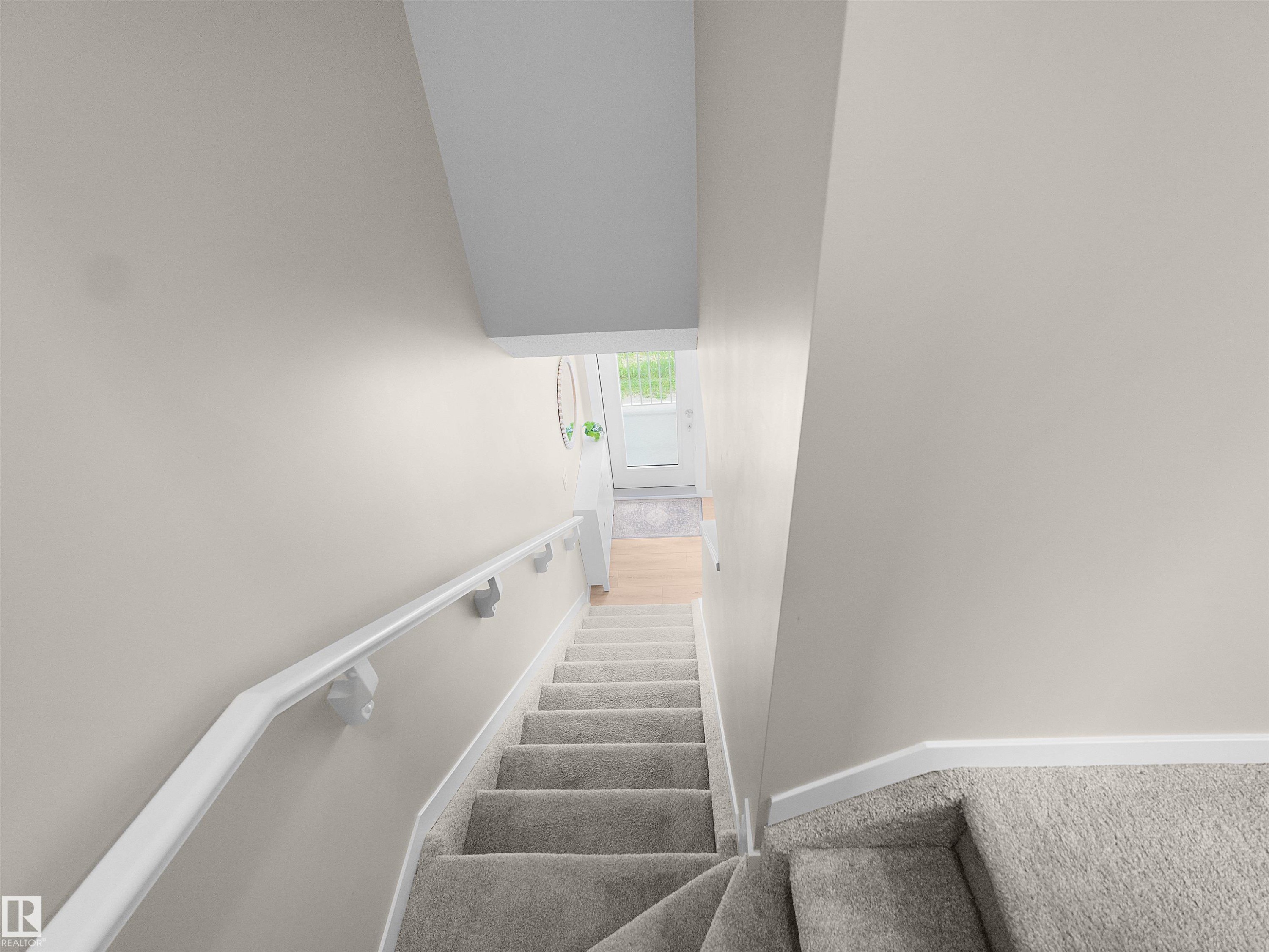 Carpeted staircase featuring a white wall-mounted handrail, neutral wall tones, white trim, and a bright entryway with a glass-paneled door - #49 17832 78 St Nw, Edmonton, AB - Indoor Photo Showing Other Room