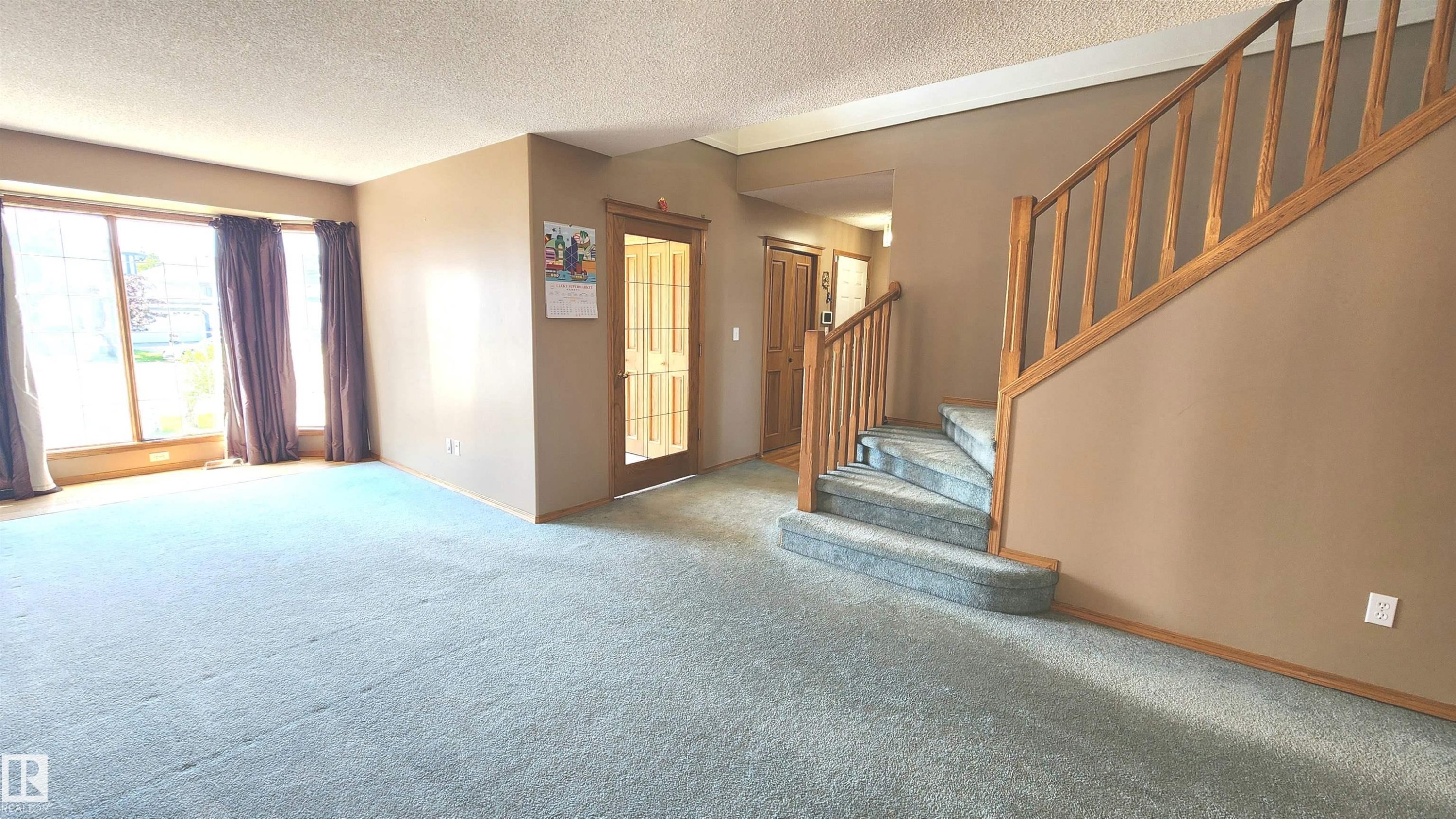 Spacious interior featuring a prominent wooden staircase with carpeted steps, large bay windows, light-colored carpeting, a front entry door with glass paneling, and an additional interior doorway - 2708 44A Avenue, Edmonton, AB