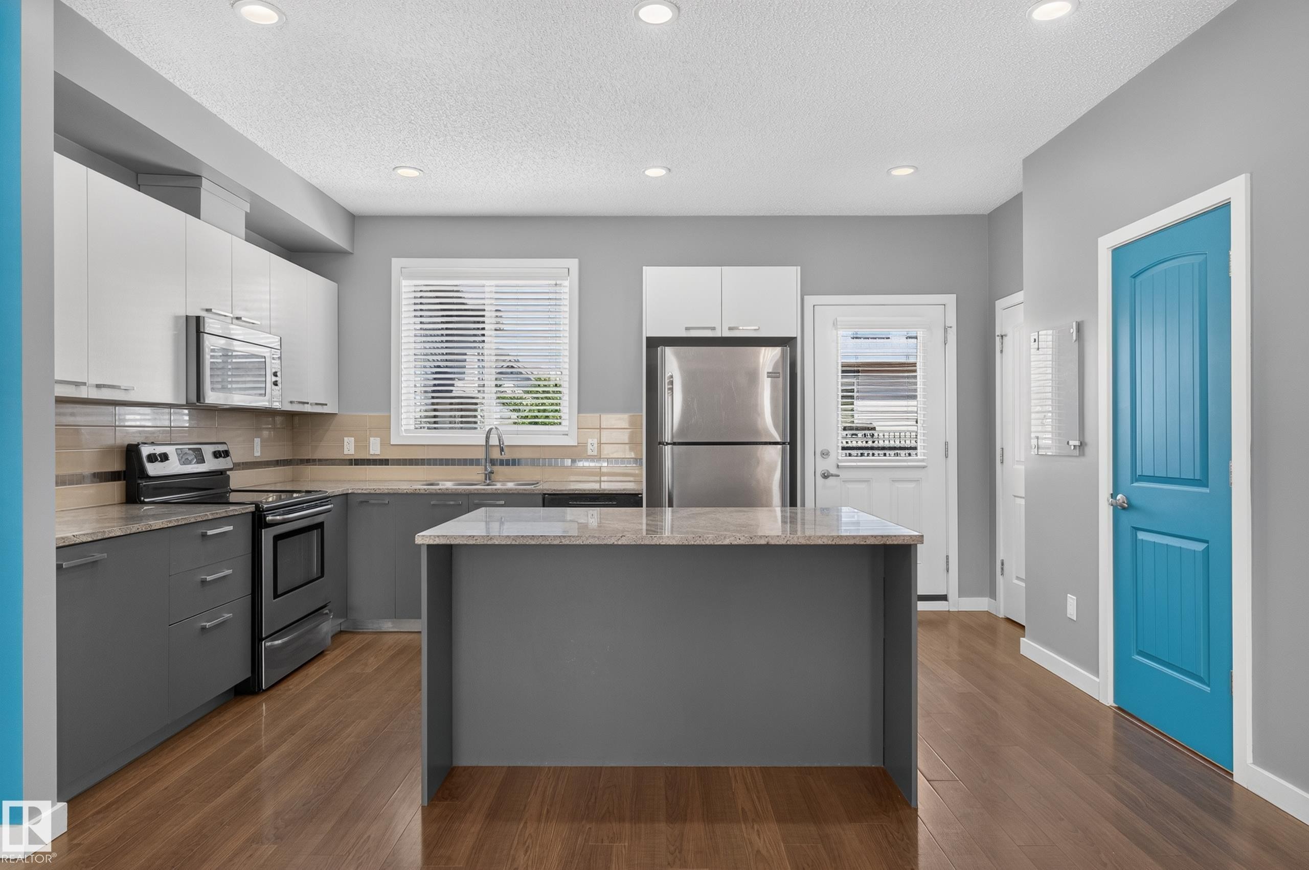 Contemporary kitchen featuring a central island, stainless steel appliances, wood-finish flooring, recessed lighting, and a vibrant blue paneled door - 24 655 Watt Boulevard, Edmonton, AB