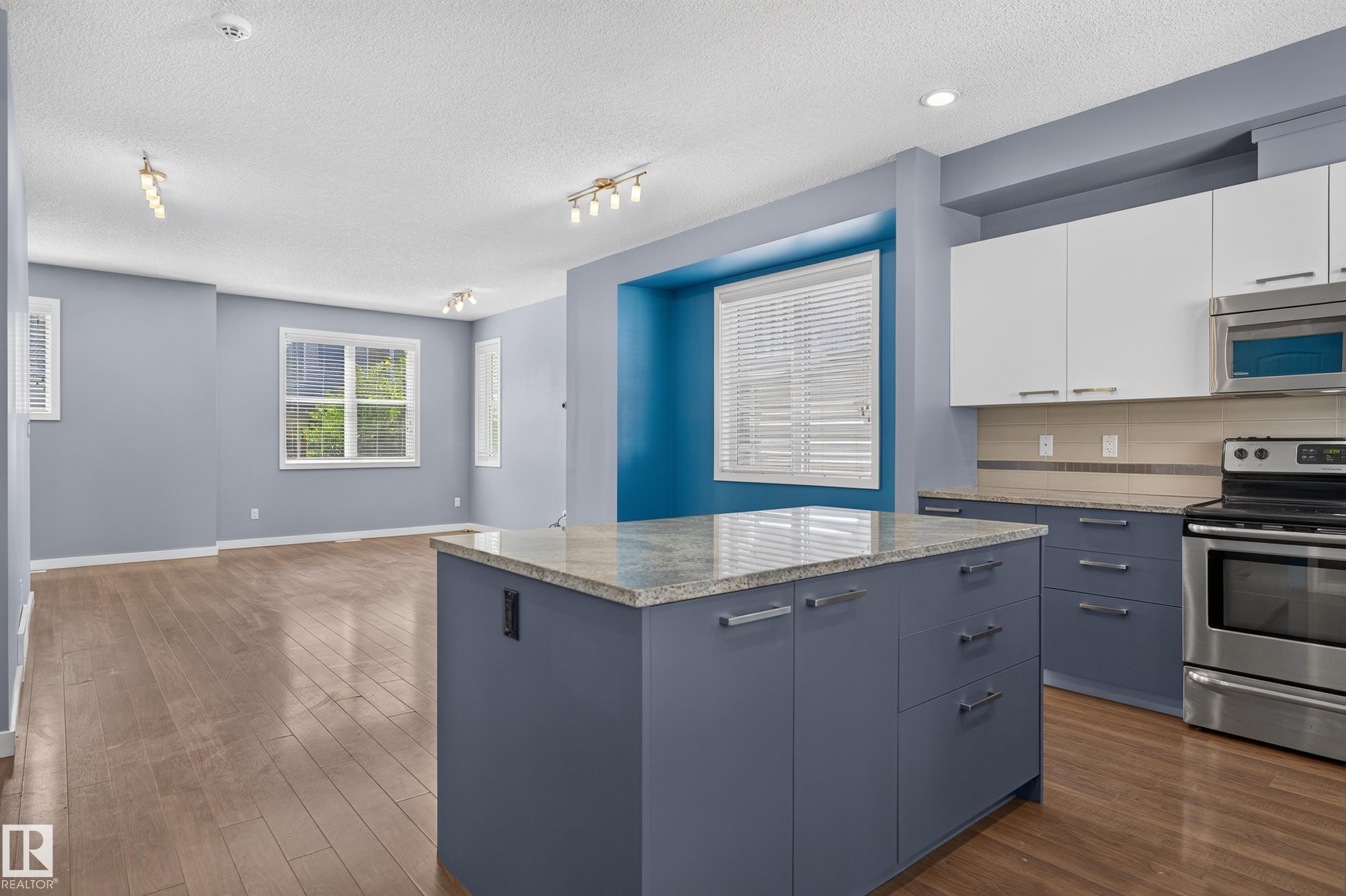 Open-concept kitchen featuring a central island with a light-toned countertop, two-tone cabinetry, stainless steel appliances, and wood-finish flooring - 24 655 Watt Boulevard, Edmonton, AB