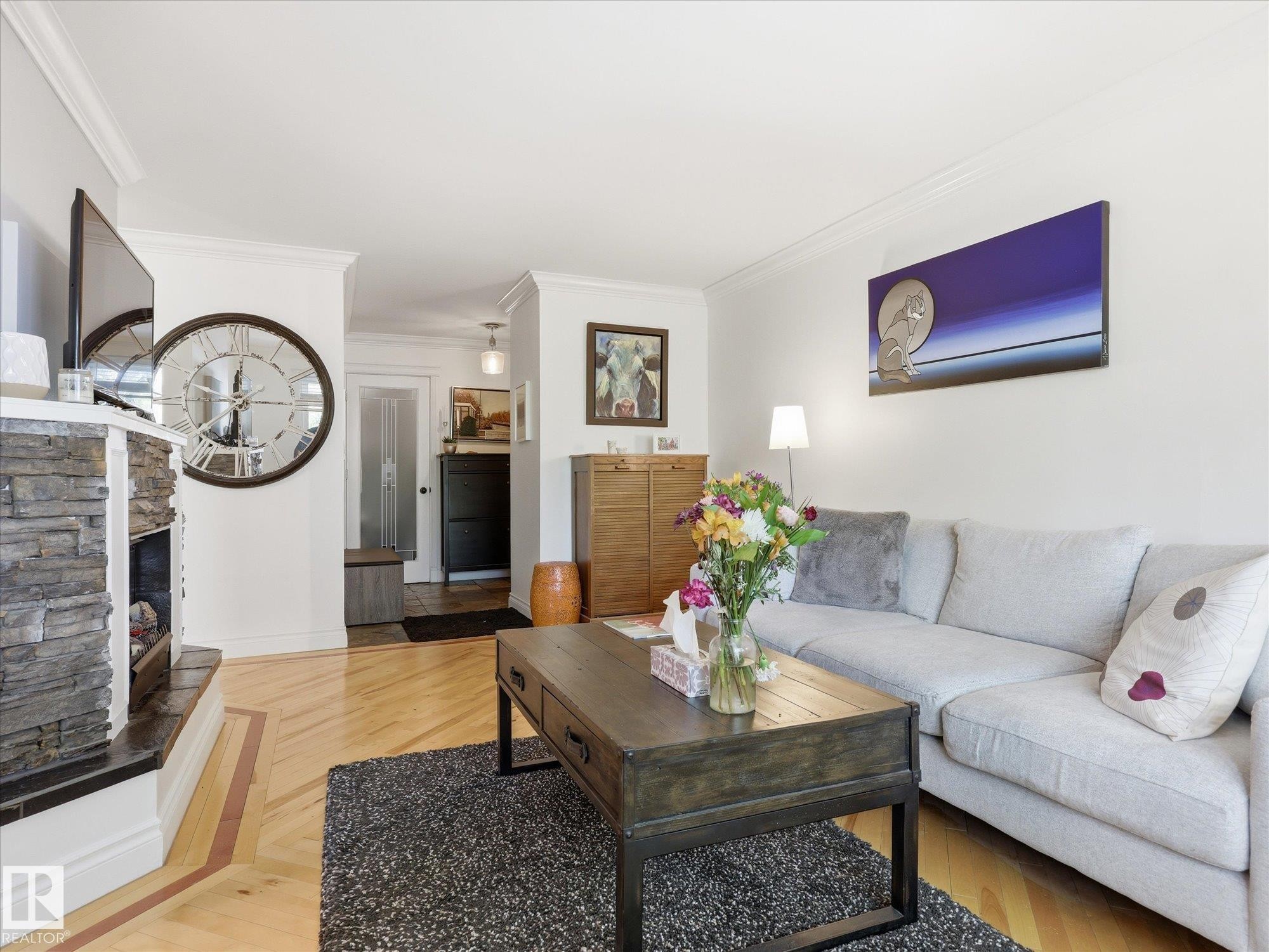 Living area featuring a stacked stone fireplace with a dark mantel, light wood-finish flooring with patterned inlay, crown molding, and white wall paint - 301 10003 87 Avenue, Edmonton, AB - Indoor Photo Showing Living Room With Fireplace