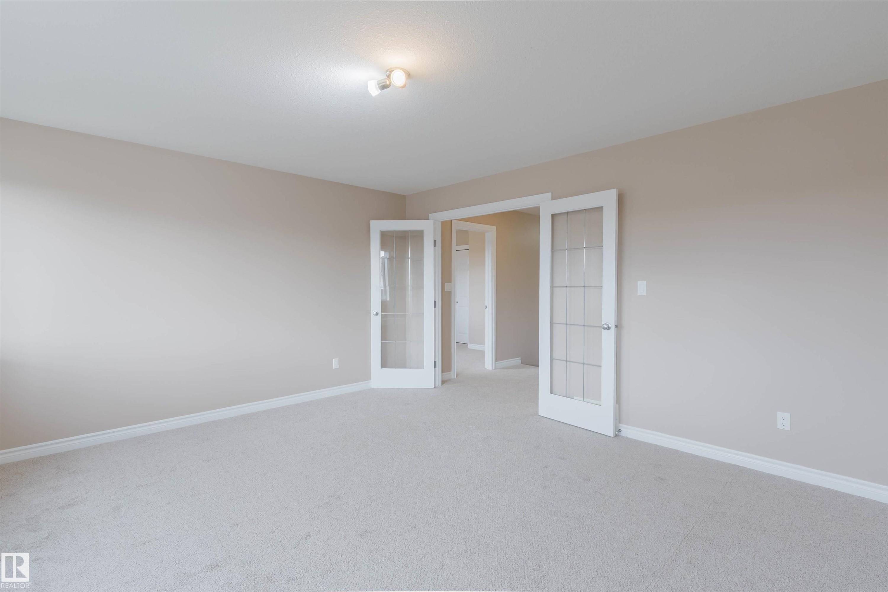 Soft neutral wall tones in this spacious room feature dual French doors with frosted glass panels, carpet flooring, and white baseboards - 1919 Hammond Place, Edmonton, AB