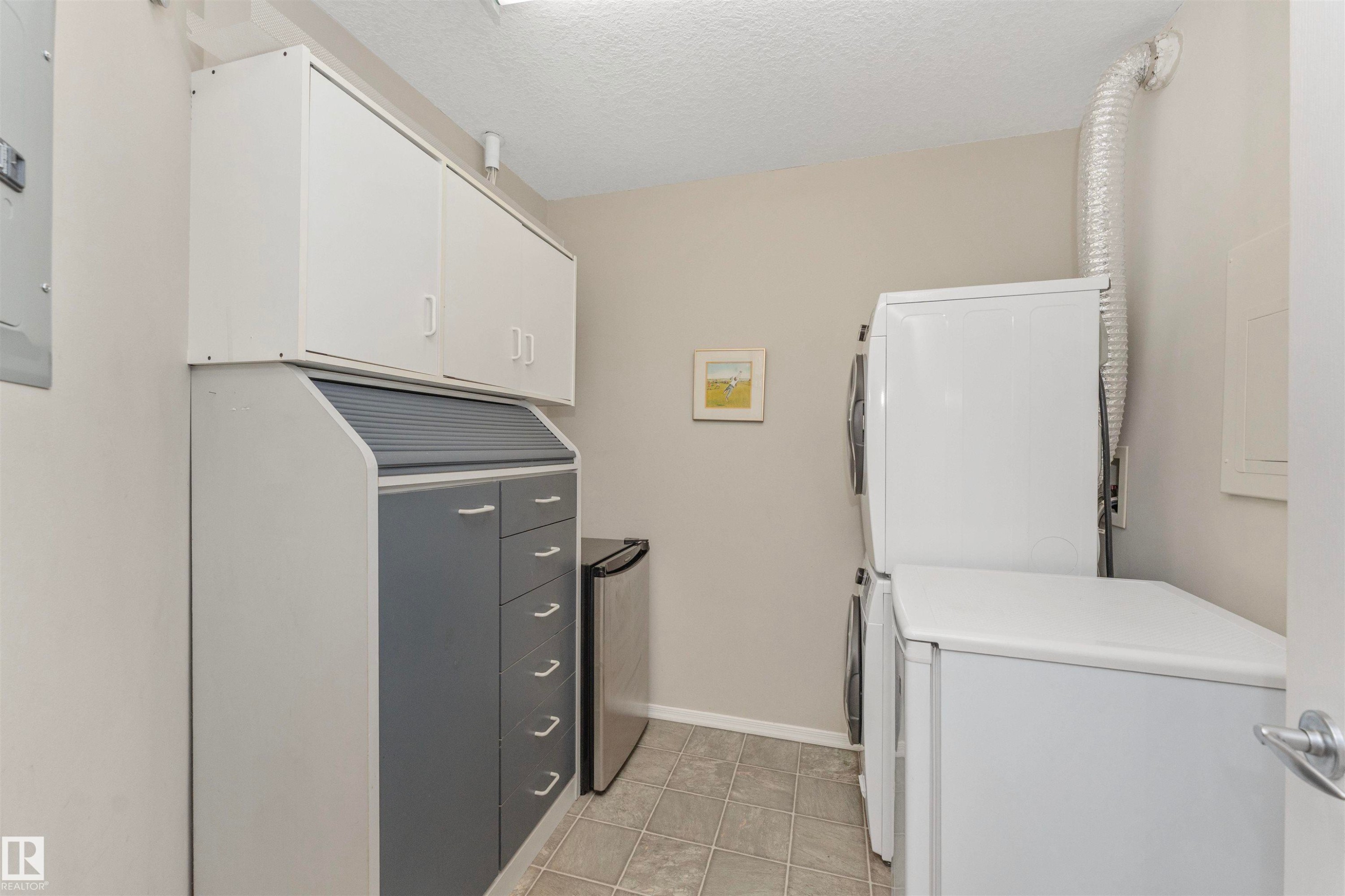 Functional utility area featuring a stacked washer and dryer, tile flooring, and ample built-in cabinetry with both white and gray finishes - #130 9008 99 Av Nw Nw, Edmonton, AB