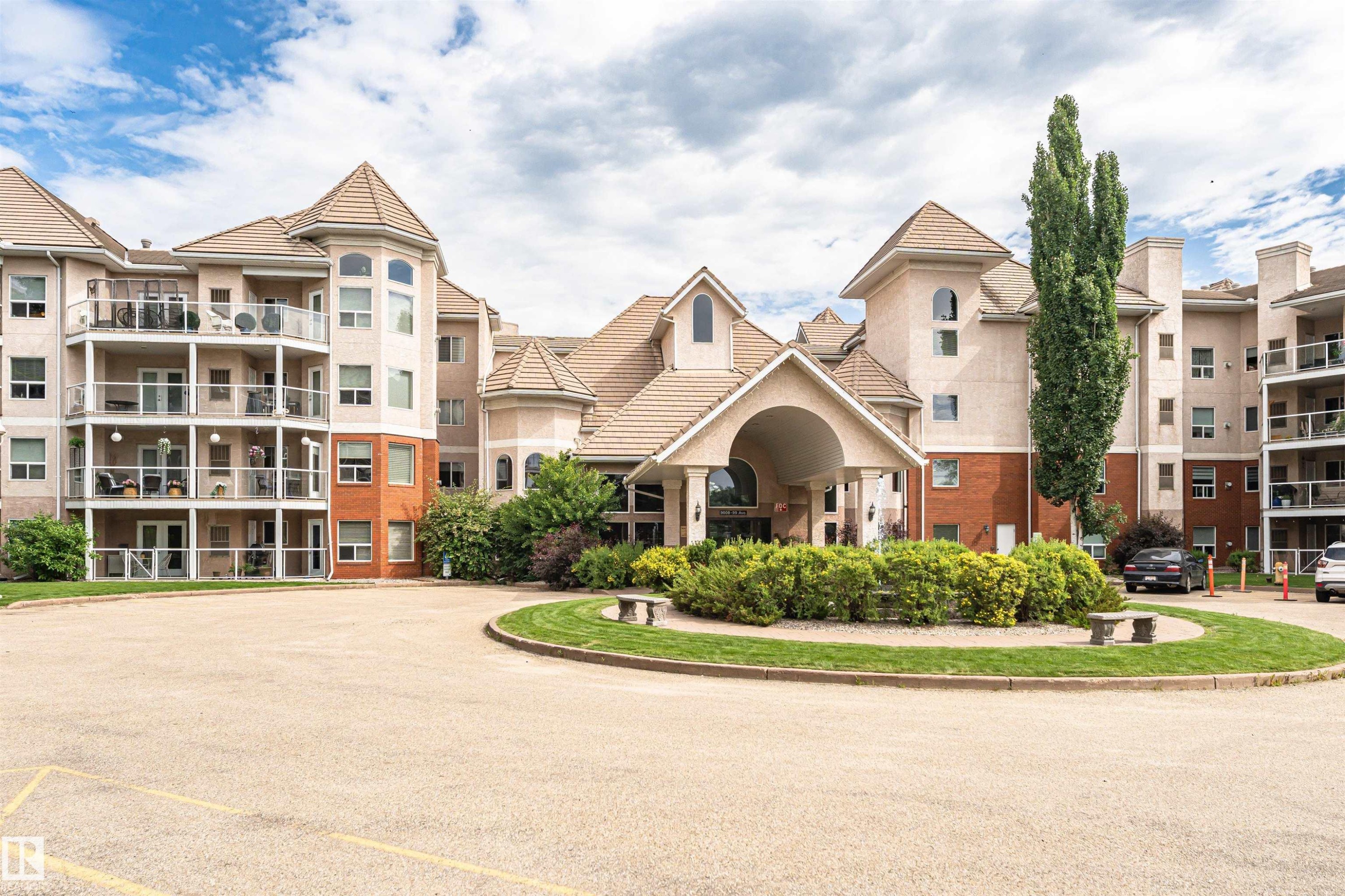 Multi-level stucco and brick exterior featuring a grand covered entrance, multiple private balconies with glass railings, and a prominent turret architectural element - #130 9008 99 Av Nw Nw, Edmonton, AB