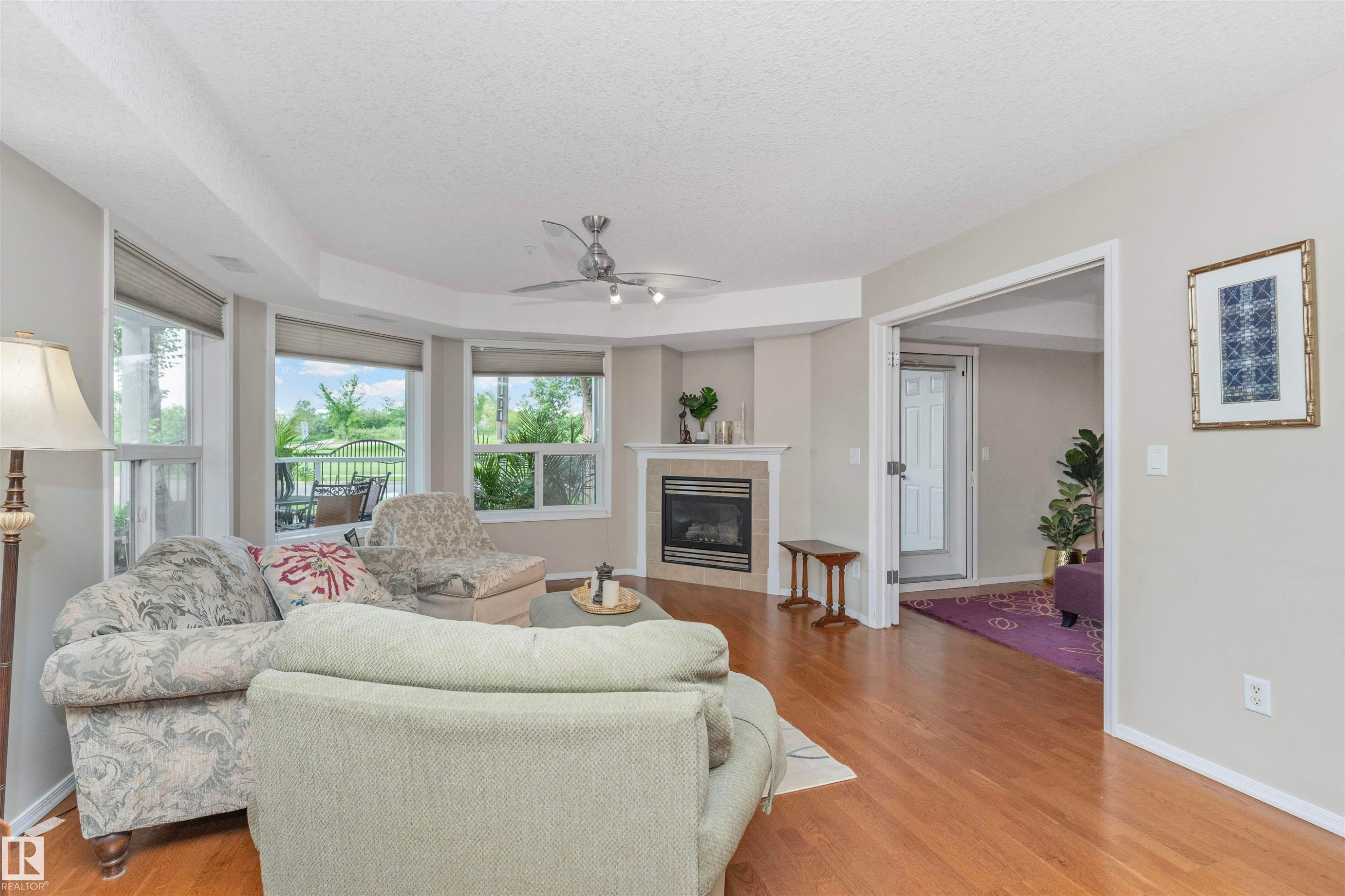 Living area featuring wood-finish flooring, a corner fireplace with tile surround, and multiple windows with roller shades - #130 9008 99 Av Nw Nw, Edmonton, AB