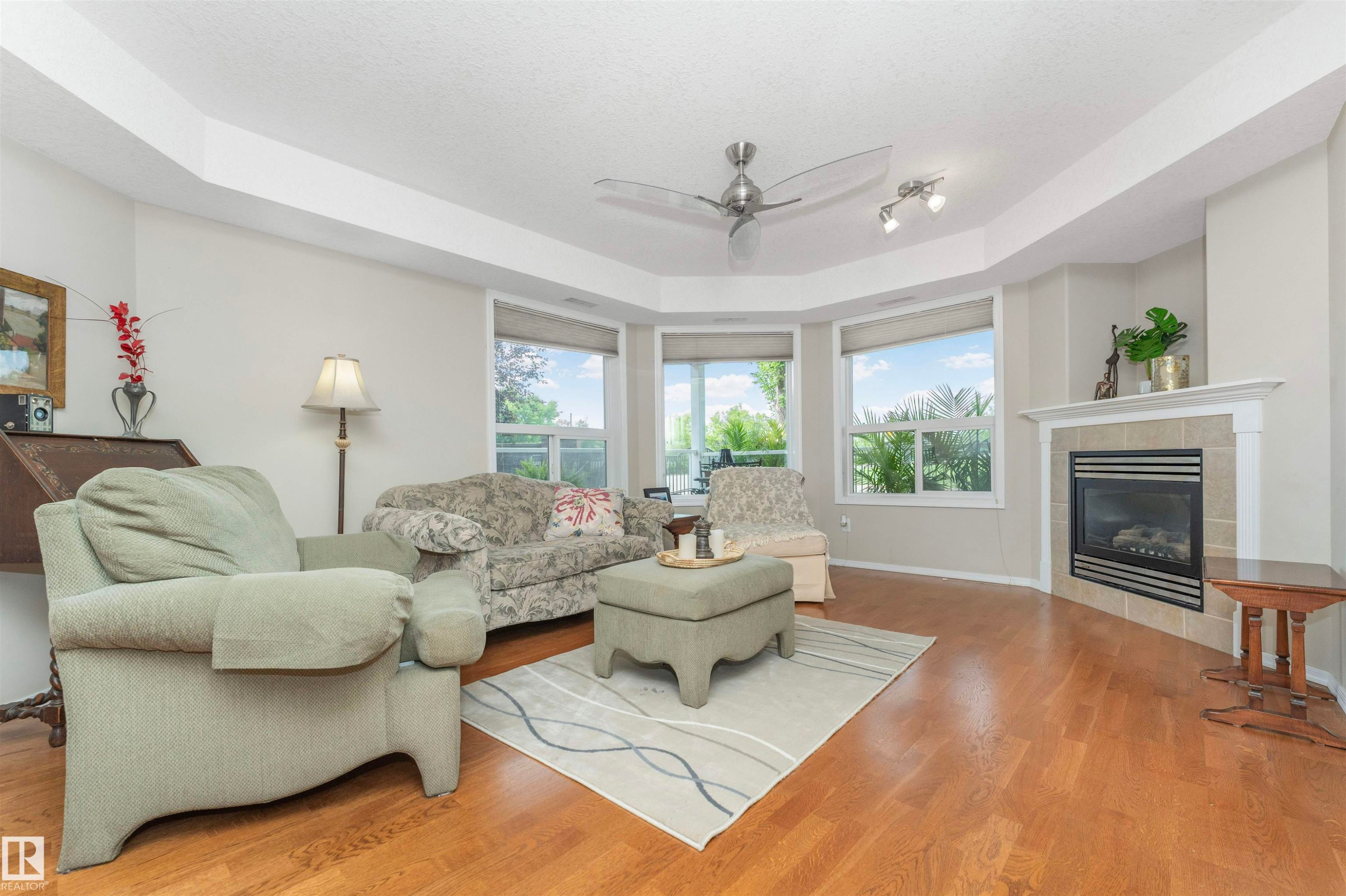 Living area with wood-finish flooring, a corner fireplace featuring a tile surround and mantel, and a bay window configuration - #130 9008 99 Av Nw Nw, Edmonton, AB