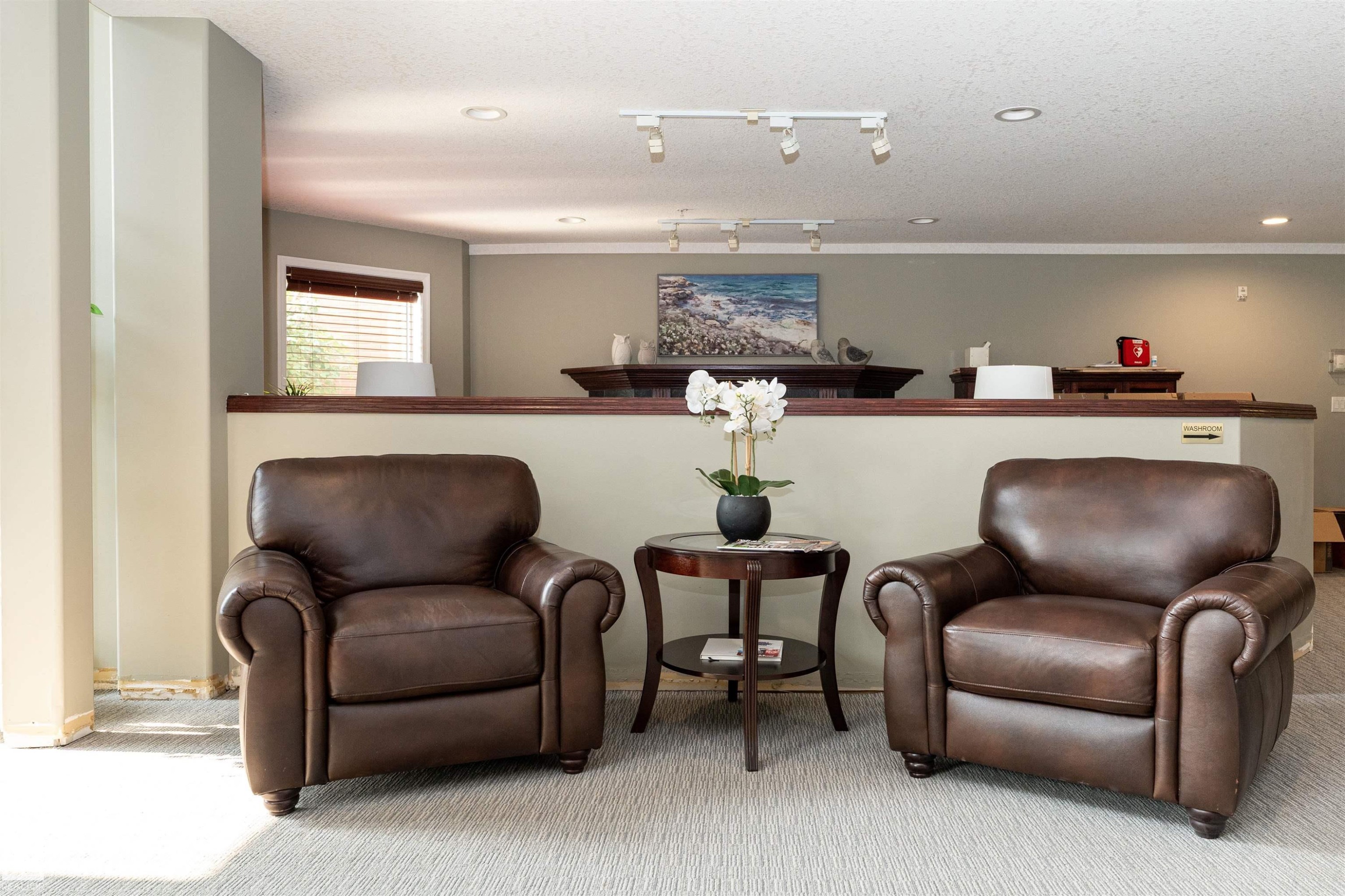 Carpeted lounge area featuring a partial wall with wood capping, track lighting, and a window with horizontal blinds - #130 9008 99 Av Nw Nw, Edmonton, AB