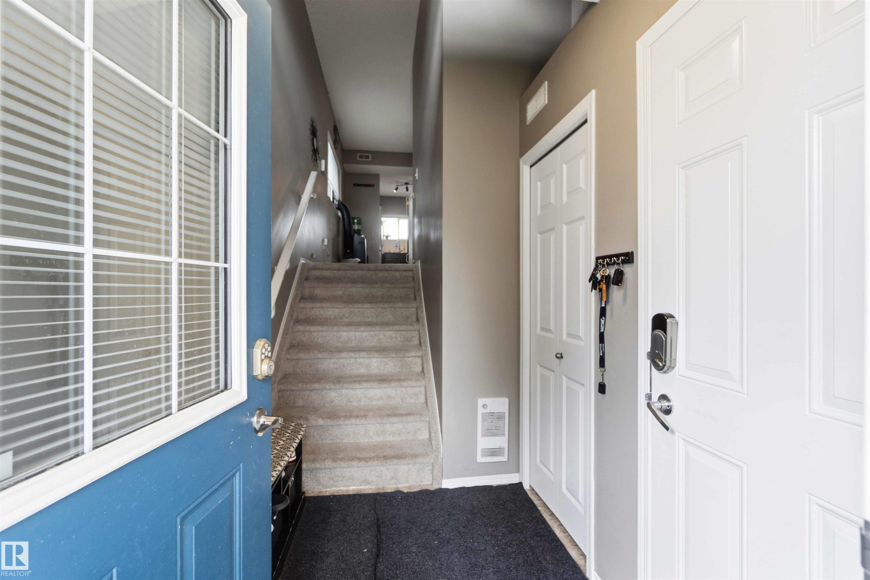 Entryway featuring a carpeted staircase, a white paneled door with hardware, a blue door with a window and white grille, and neutral wall tones - 24 6520 2 Avenue, Edmonton, AB