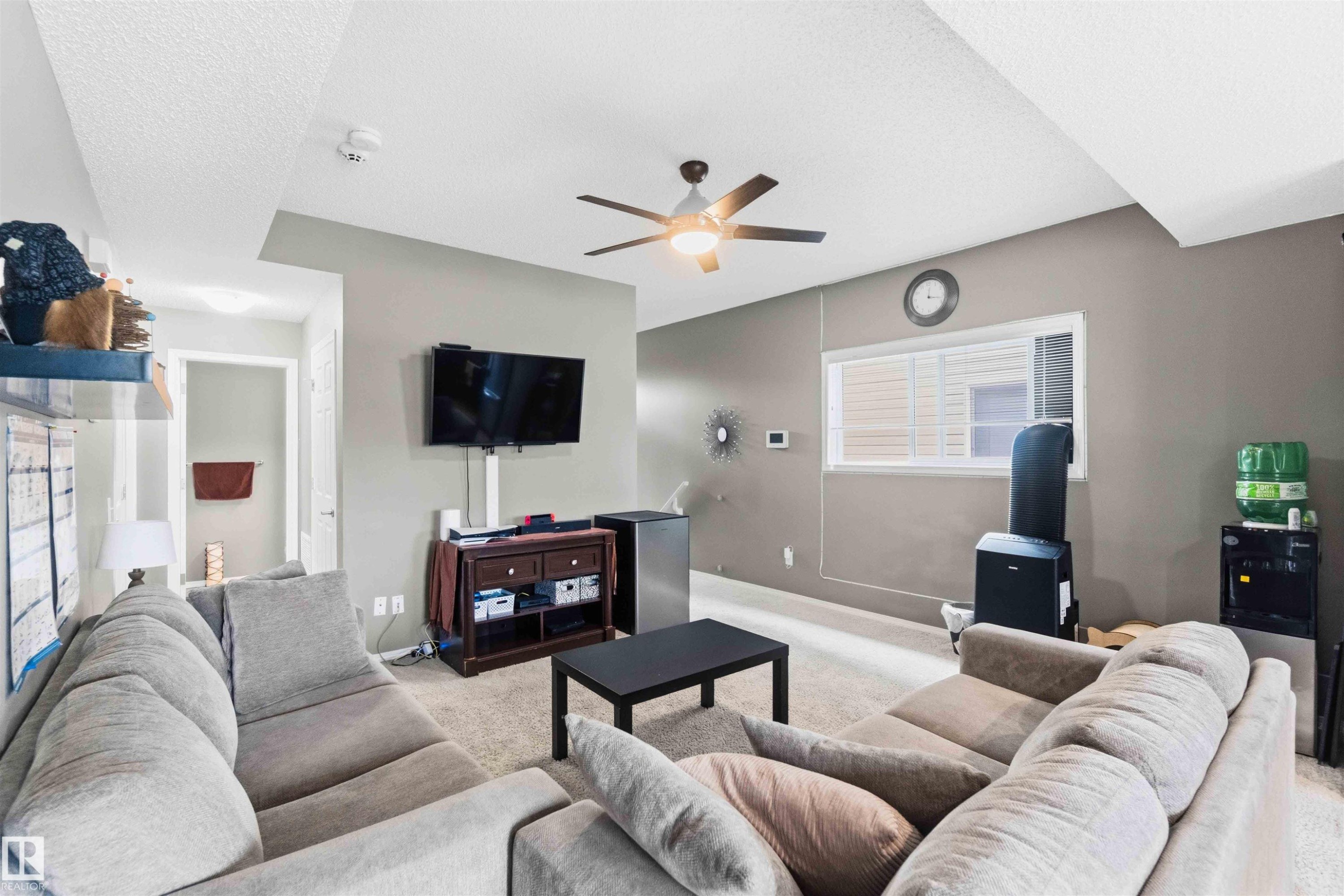 Carpeted living area featuring a ceiling fan with integrated lighting, a wall-mounted television, and a window with horizontal blinds - 24 6520 2 Avenue, Edmonton, AB