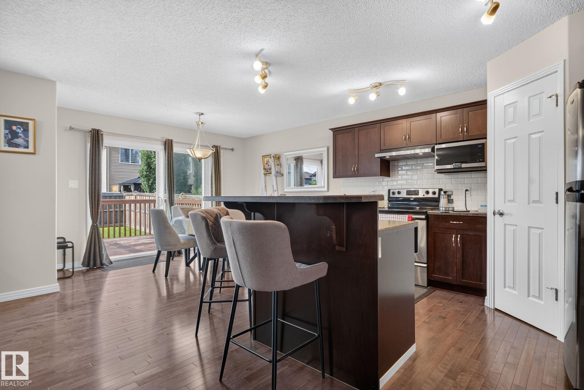 Open-concept kitchen and dining area featuring rich wood-finish flooring, dark wood cabinetry, stainless steel appliances, a white subway tile backsplash, and a breakfast bar with seating - 7858 170A Avenue, Edmonton, AB