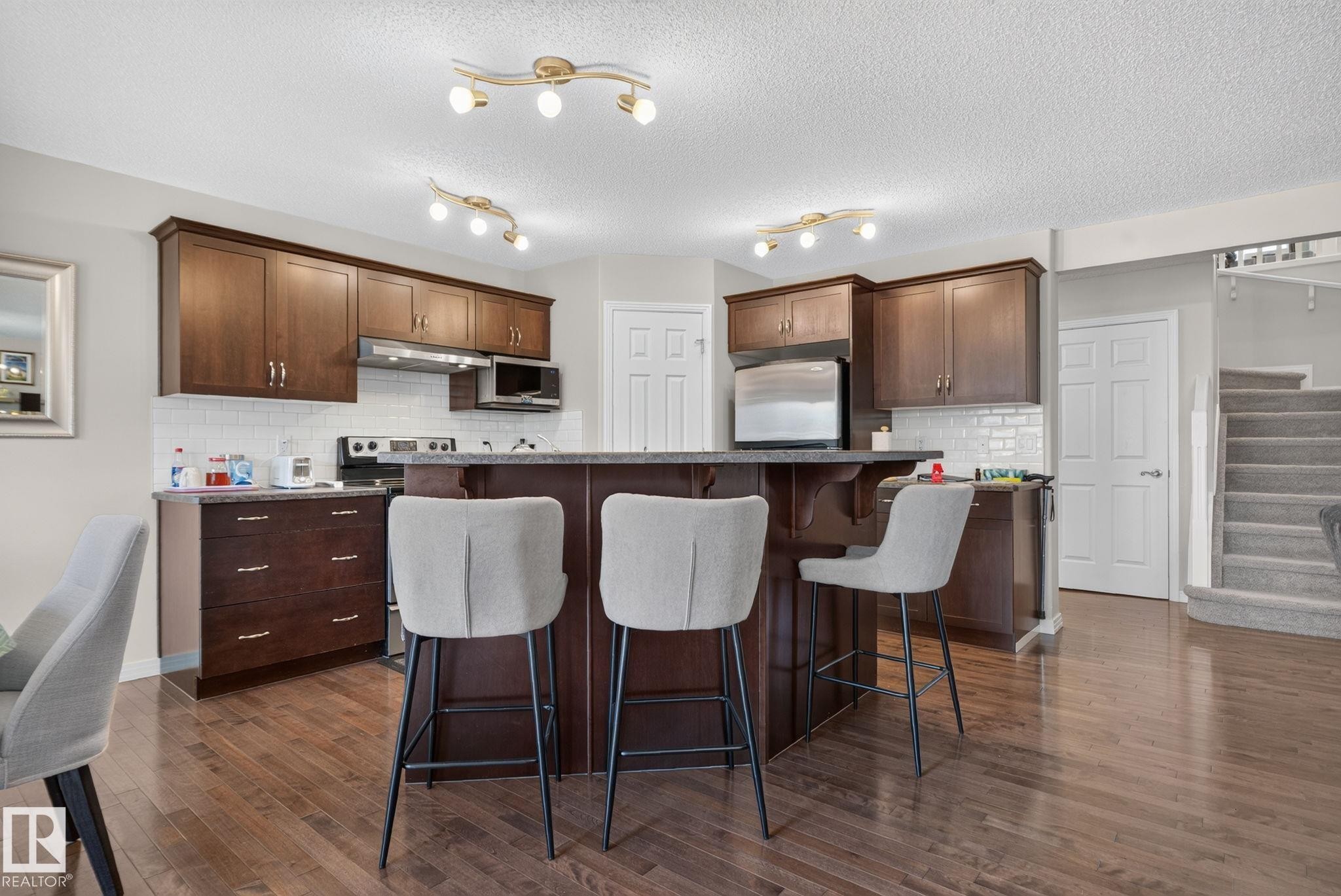 Kitchen featuring wood-finish flooring, dark wood cabinetry, a central island with bar seating, stainless steel appliances, and a white subway tile backsplash - 7858 170A Avenue, Edmonton, AB