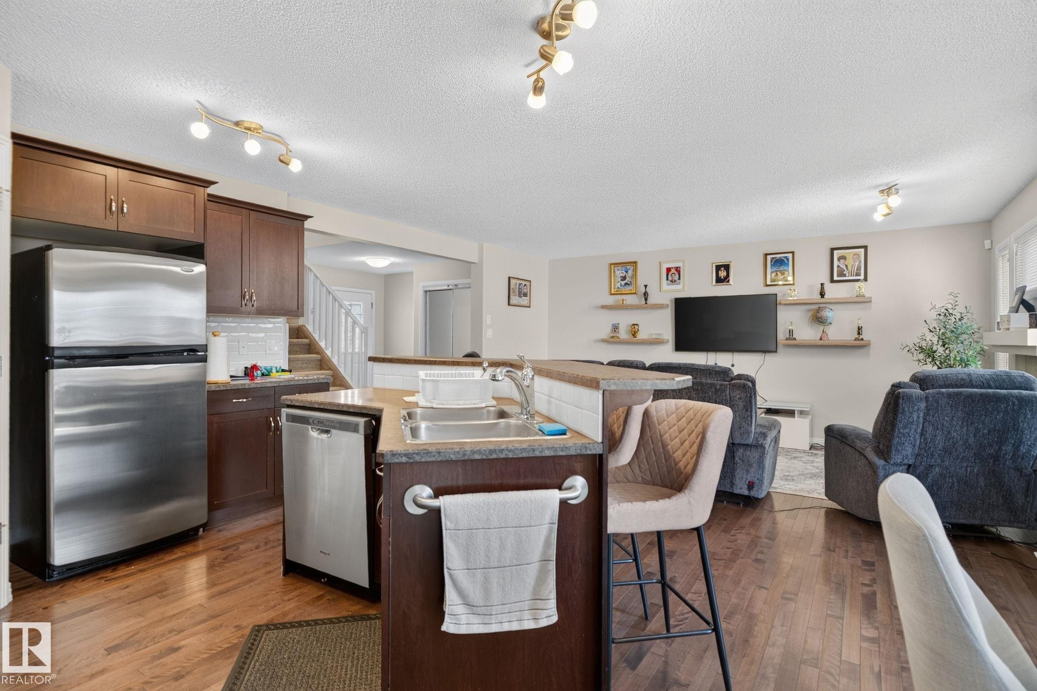 Open-concept kitchen featuring wood-finish flooring, stainless steel appliances, dark wood cabinetry, and a tiered island with integrated sink - 7858 170A Avenue, Edmonton, AB