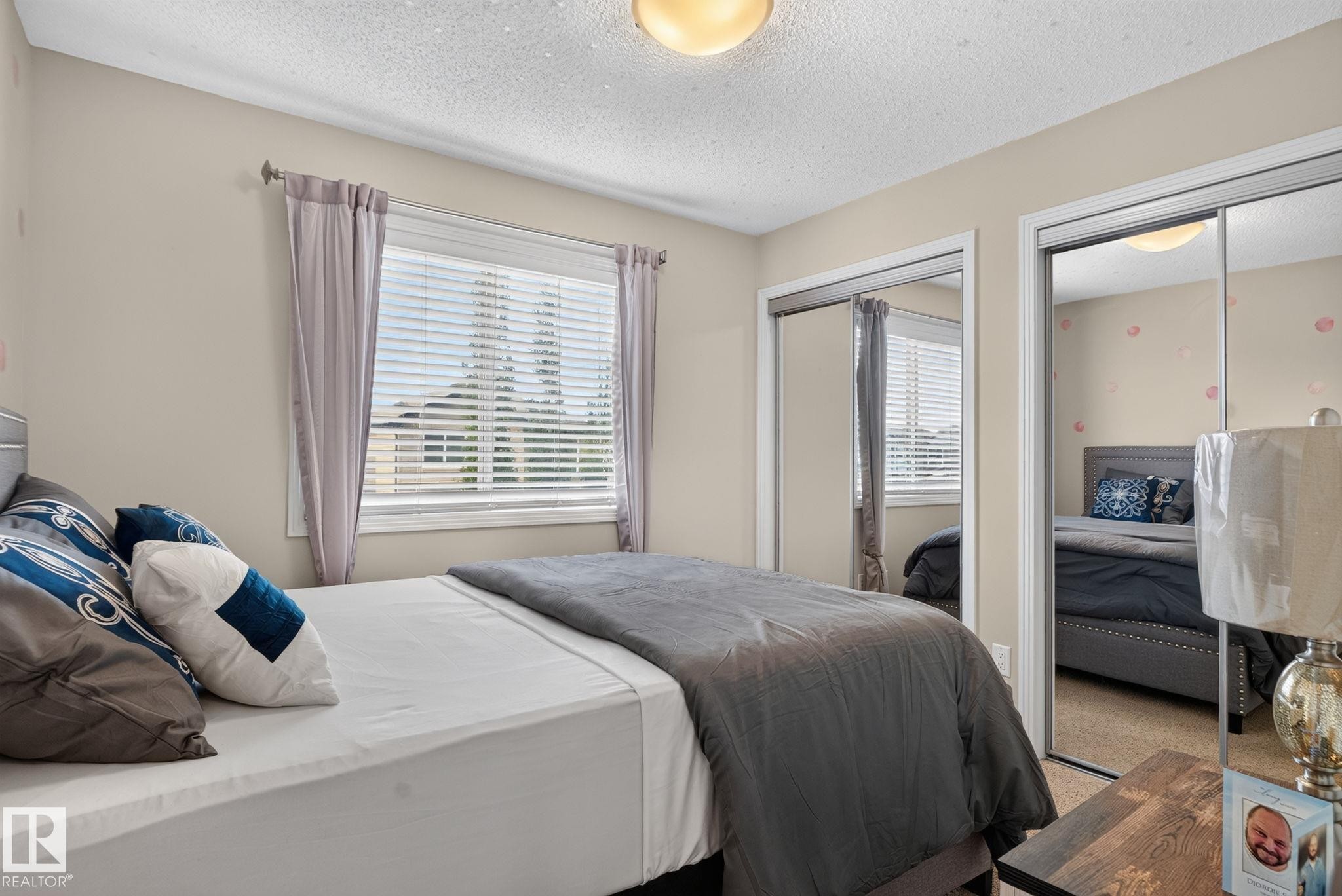 Bedroom featuring a window with horizontal blinds and curtains, two mirrored sliding closet doors, a flush-mount ceiling light, and neutral wall paint - 7858 170A Avenue, Edmonton, AB