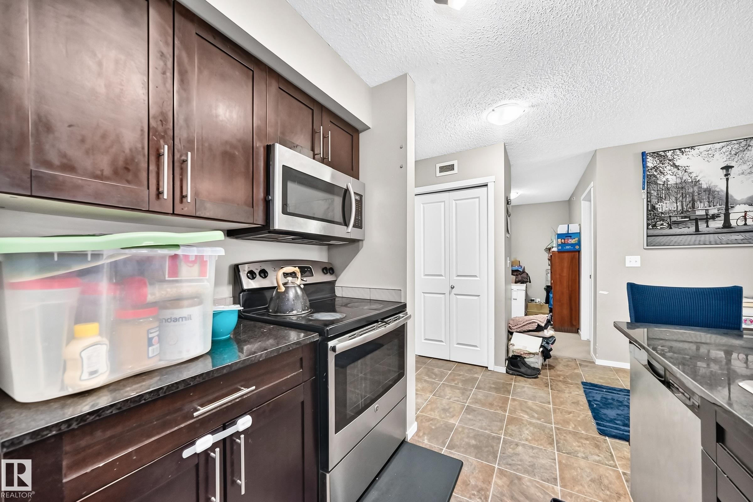 Kitchen featuring dark wood cabinetry, stainless steel appliances including a microwave and range, dark stone countertops, and square floor tiles - #417 1510 Watt Dr Sw, Edmonton, AB - Indoor Photo Showing Kitchen